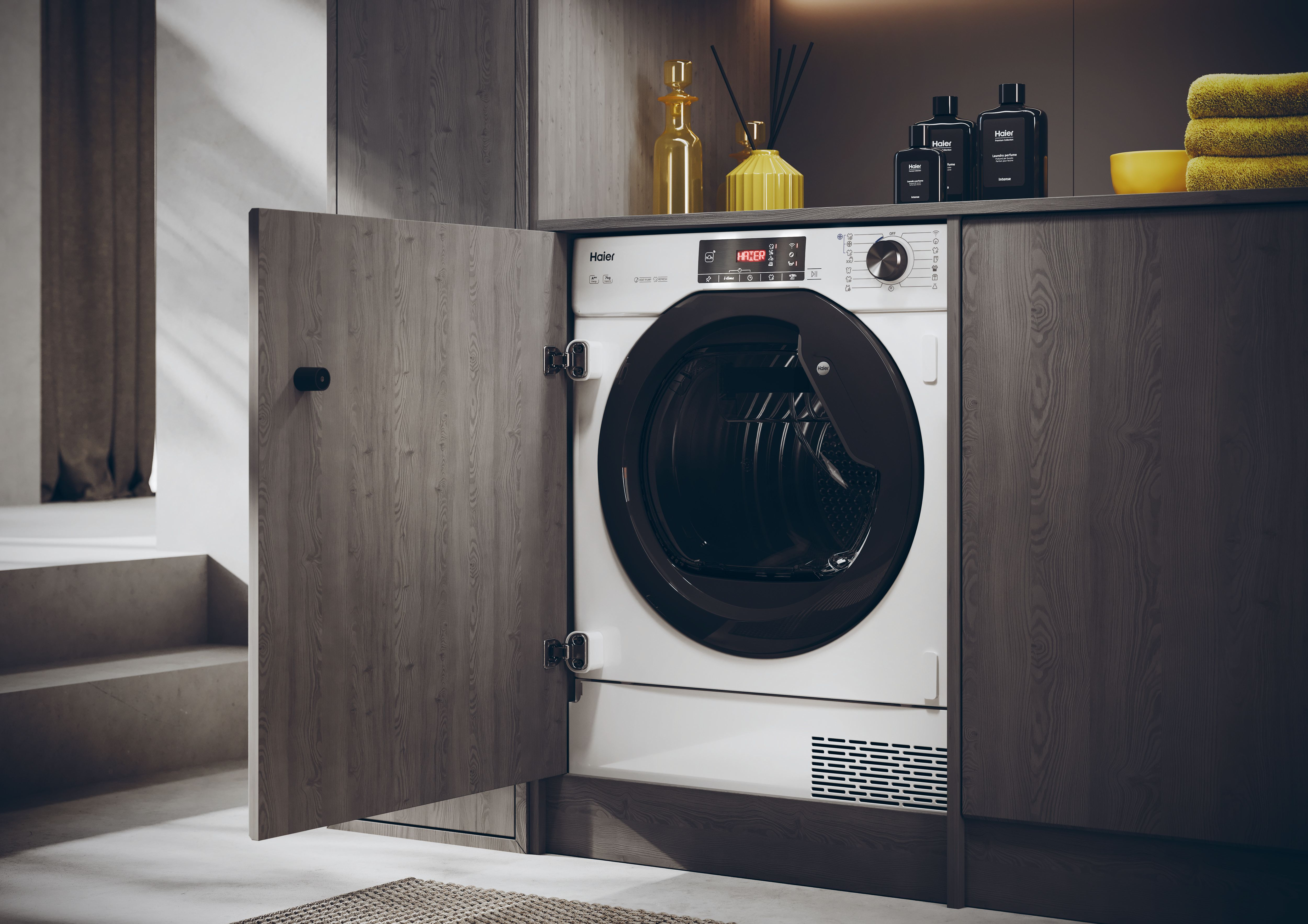 Biała suszarka Haier z otwartymi drzwiami, w drewnianej szafce. Butelki i żółte ręczniki na półce.