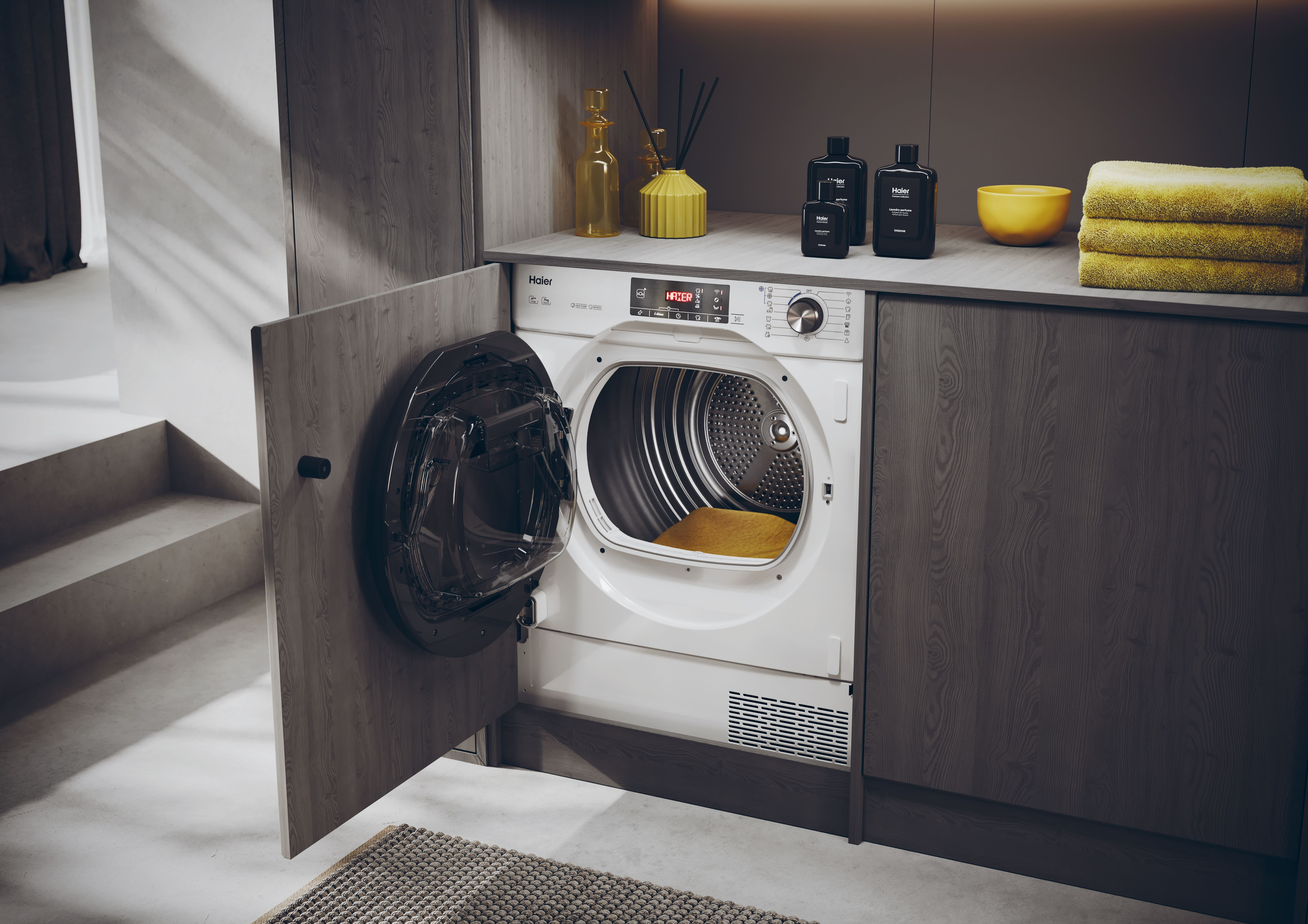 Biała suszarka Haier z otwartymi drzwiami. Na blacie znajdują się żółte ręczniki, butelki i miska.