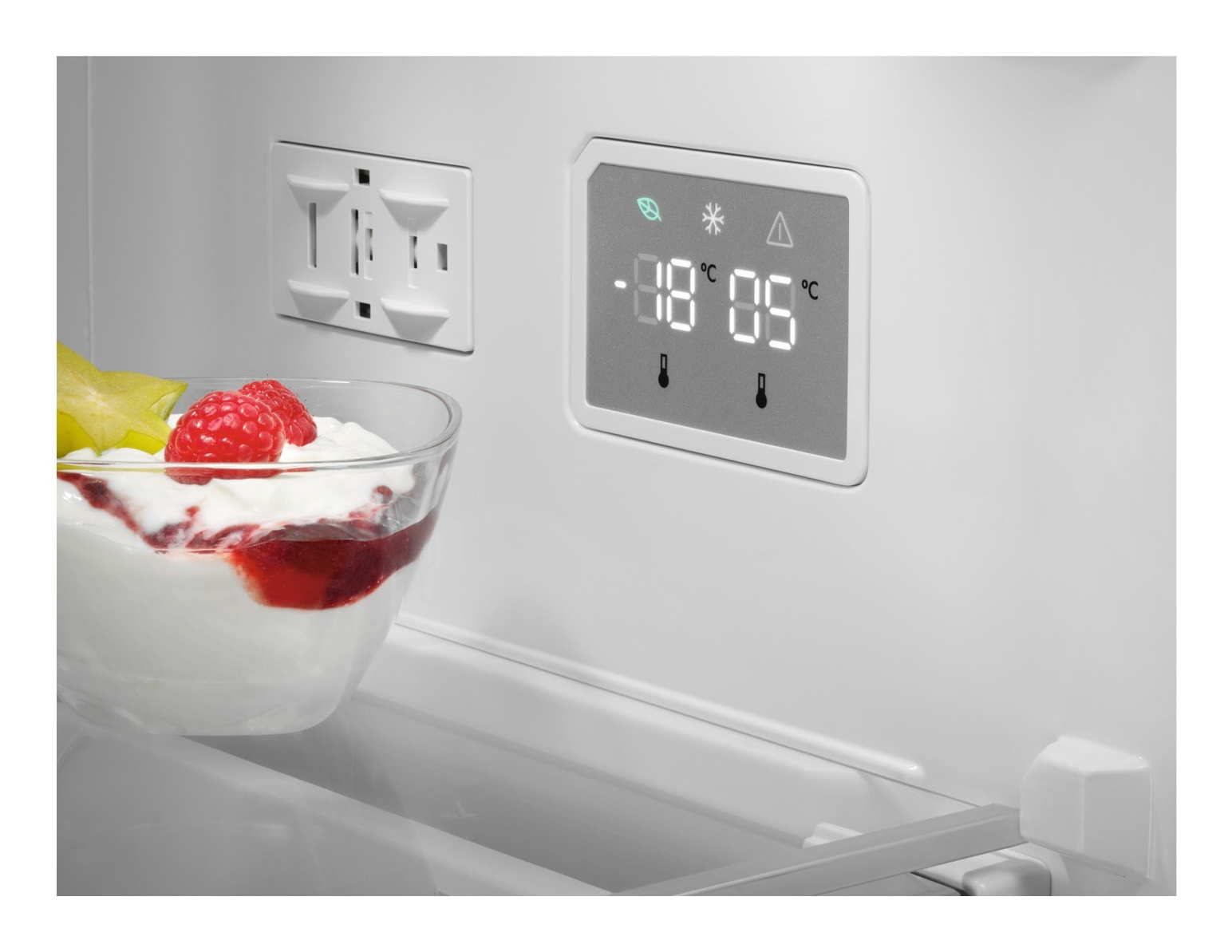 Dentro de un refrigerador, se ve un postre con fruta. Una pantalla digital muestra la temperatura. Hay un enchufe en la pared.