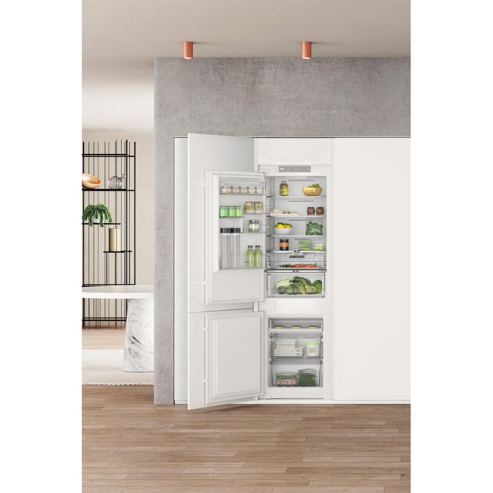 Refrigerador blanco abierto lleno de comida. Suelo de madera y pared gris al fondo. Dos luces en el techo.