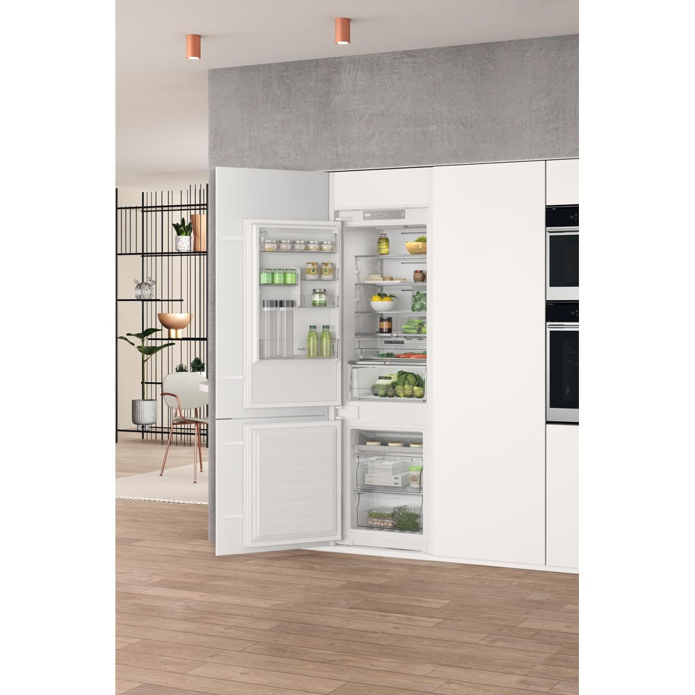 Cocina moderna con nevera blanca grande abierta, llena de comida. Suelo de madera, pared gris y dos lámparas naranjas en el techo.