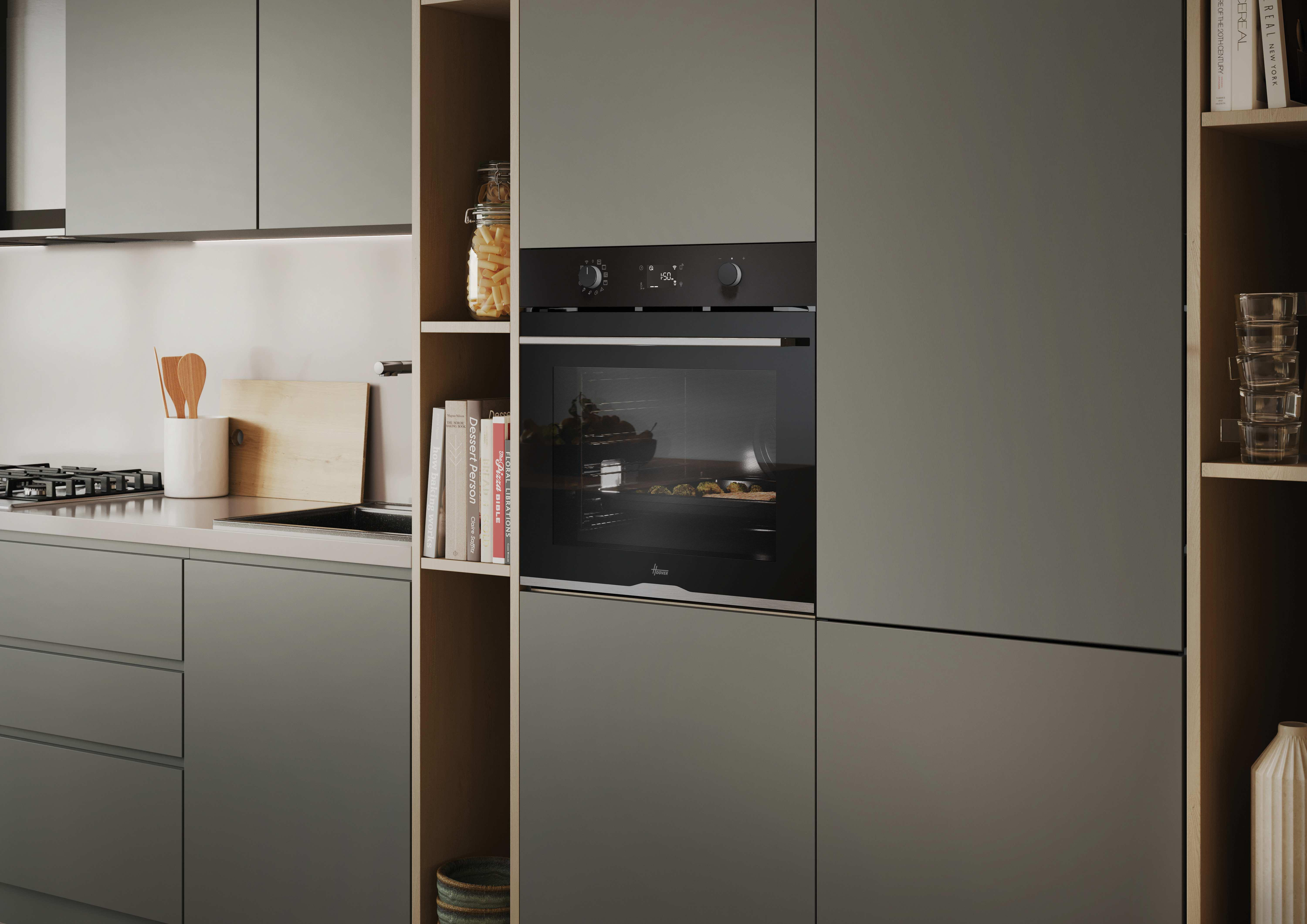 Una cucina moderna con forno nero, ripiani con libri e barattoli. Sono visibili mobili grigi e un tagliere in legno.