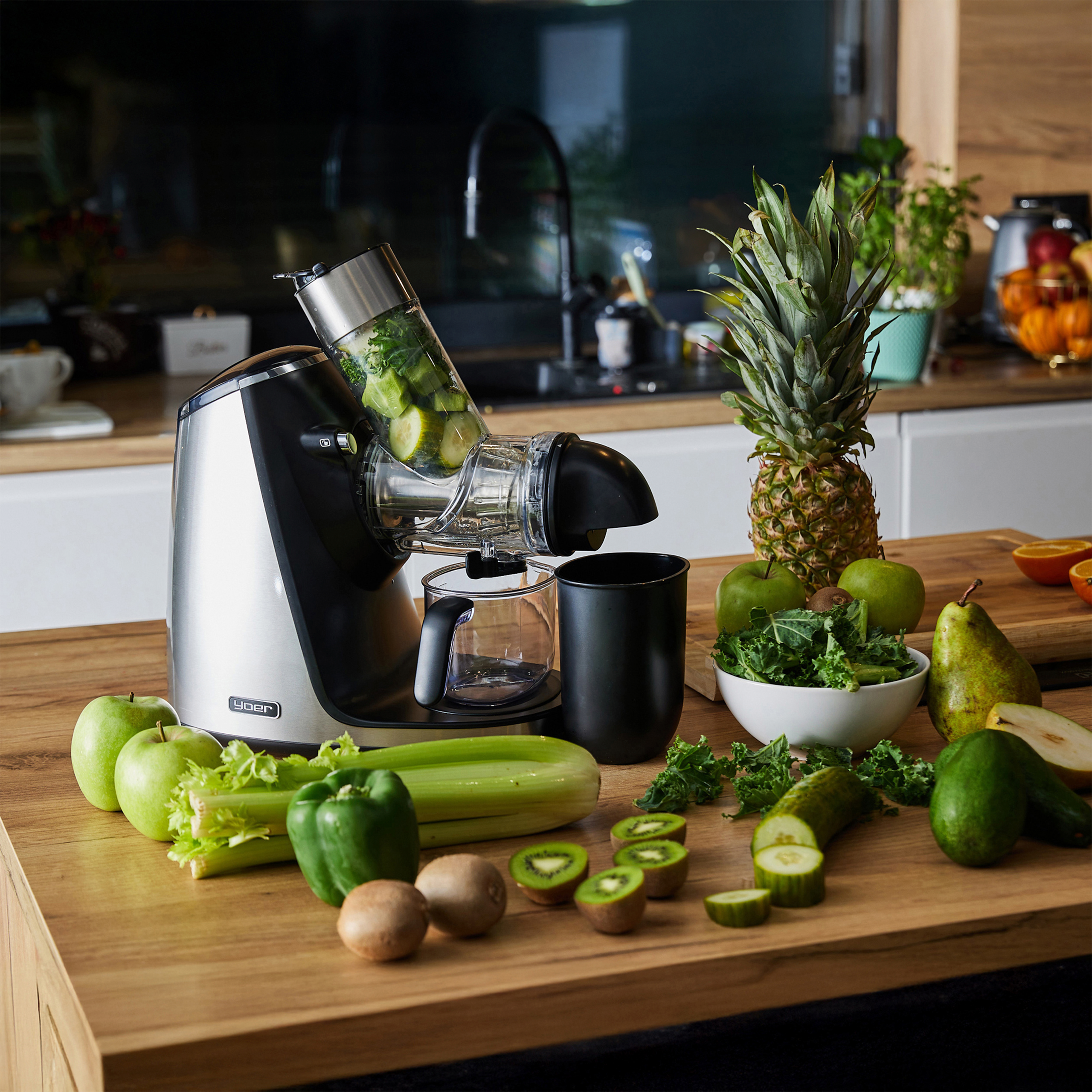 Wyrażacz soku z owocami i warzywami na drewnianym blacie. Widoczne ananasy, jabłka, gruszki, seler, ogórek i kiwi.