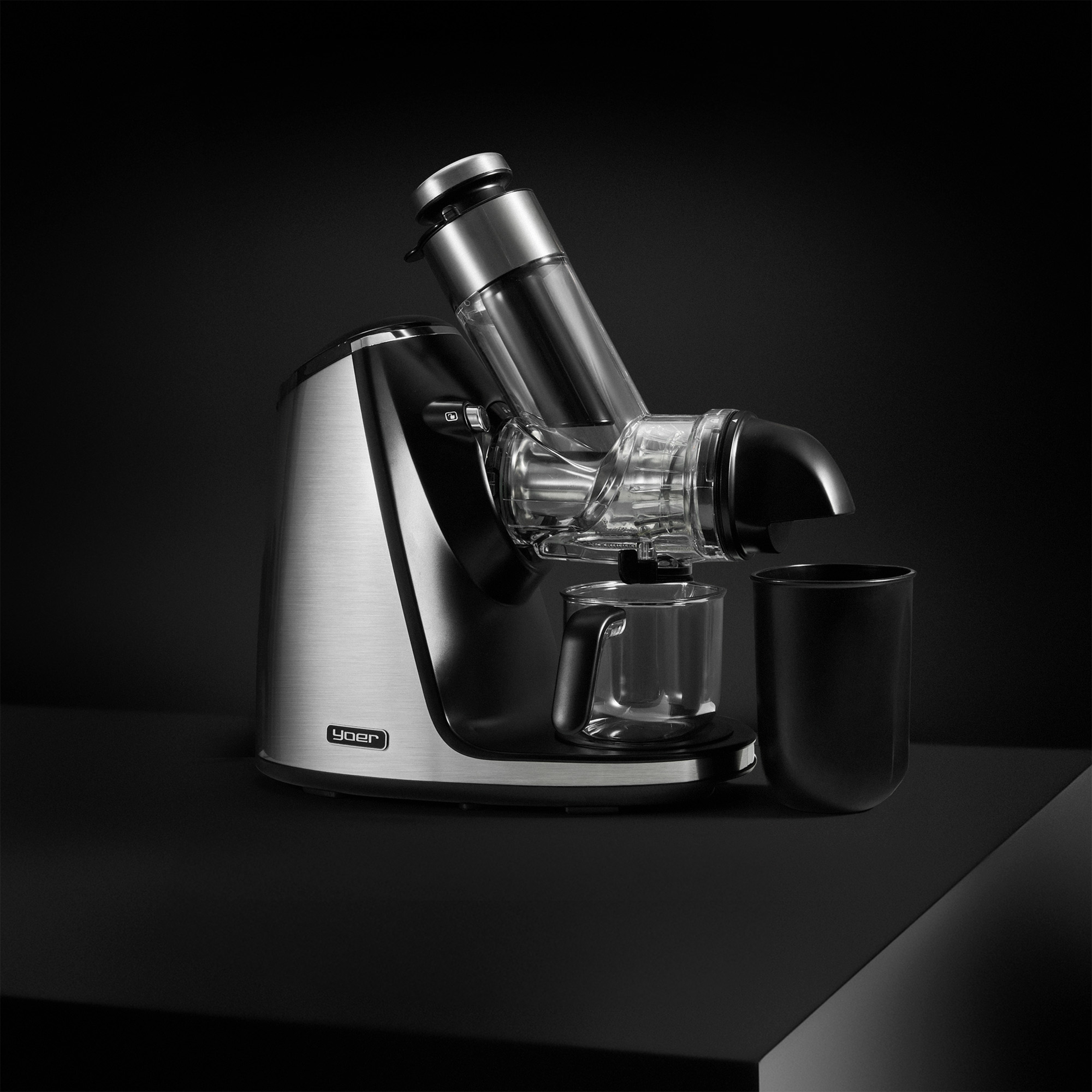 Nowoczesna wyciskarka do soków w srebrno-czarnym designie na czarnej powierzchni. Obok szklanka i czarny pojemnik.