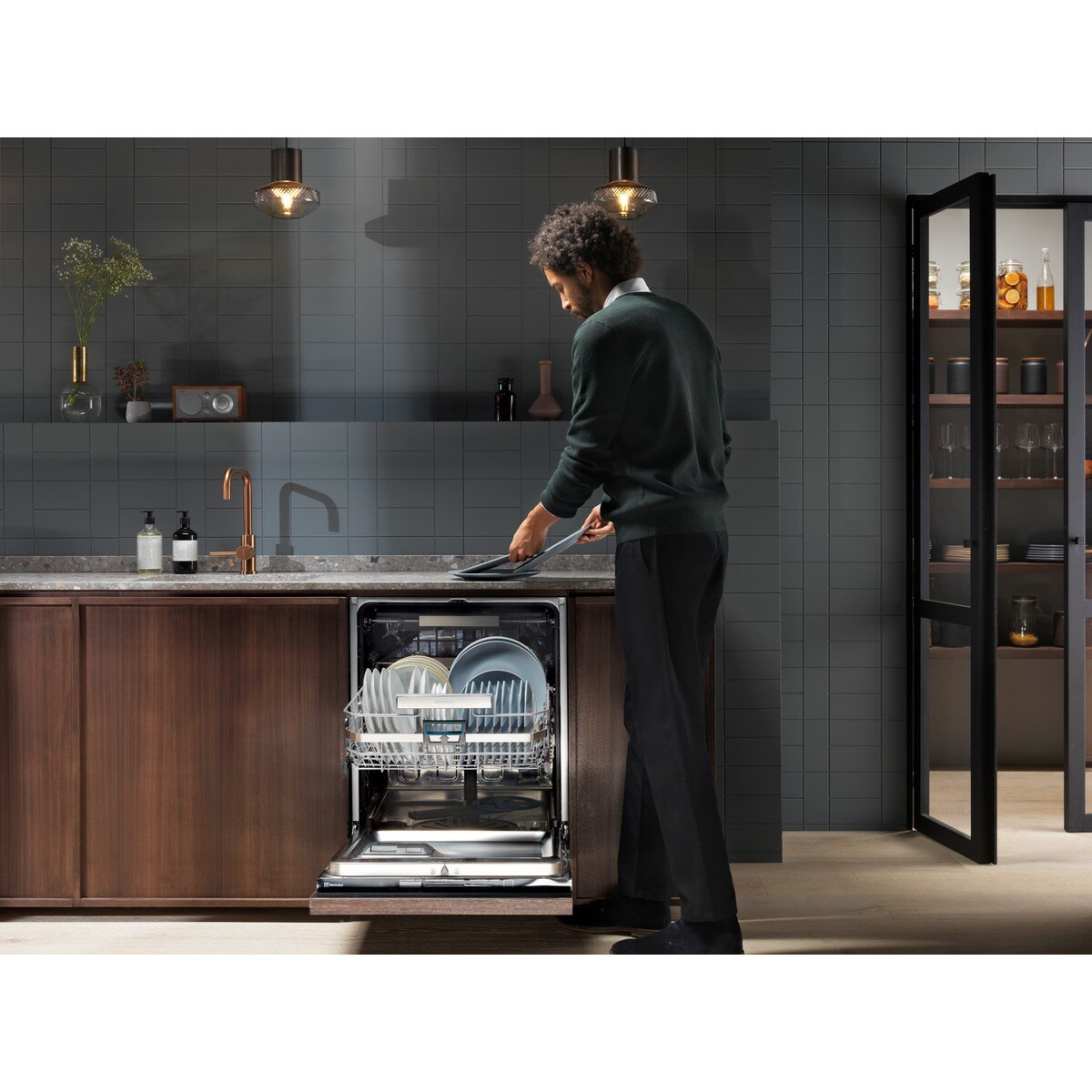 Un uomo con un maglione verde carica una lavastoviglie in una cucina moderna. Piatti all'interno e un armadio a destra.