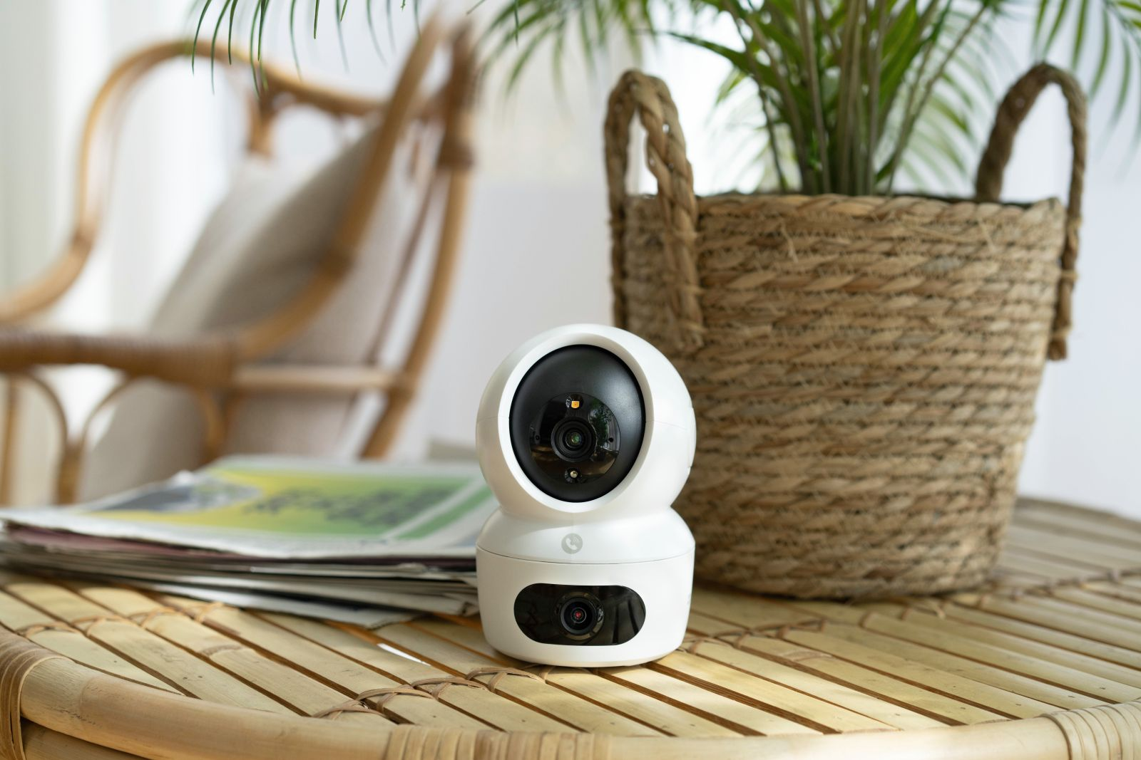 Biała kamera bezpieczeństwa stoi na bambusowym stole. Pleciony kosz z rośliną i wiklinowe krzesło są w tle.