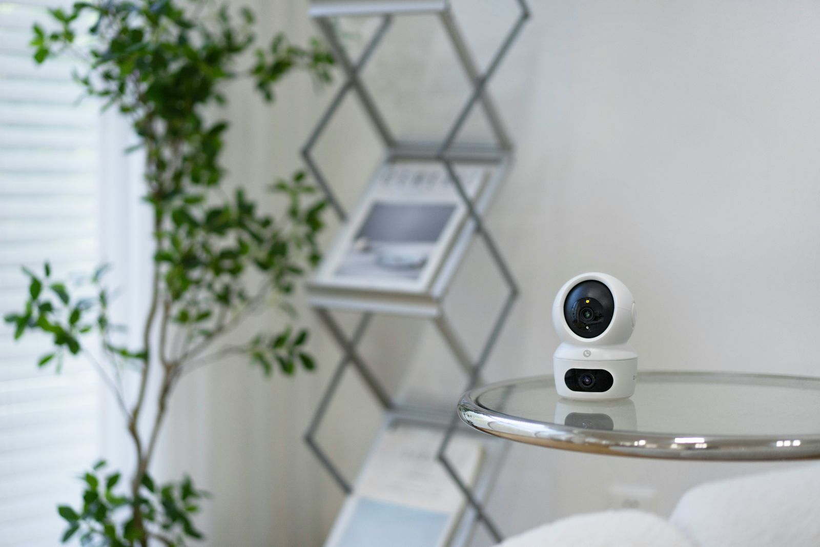 Biała kamera bezpieczeństwa stoi na szklanym stoliku. Zielona roślina i dekoracyjna półka w tle.