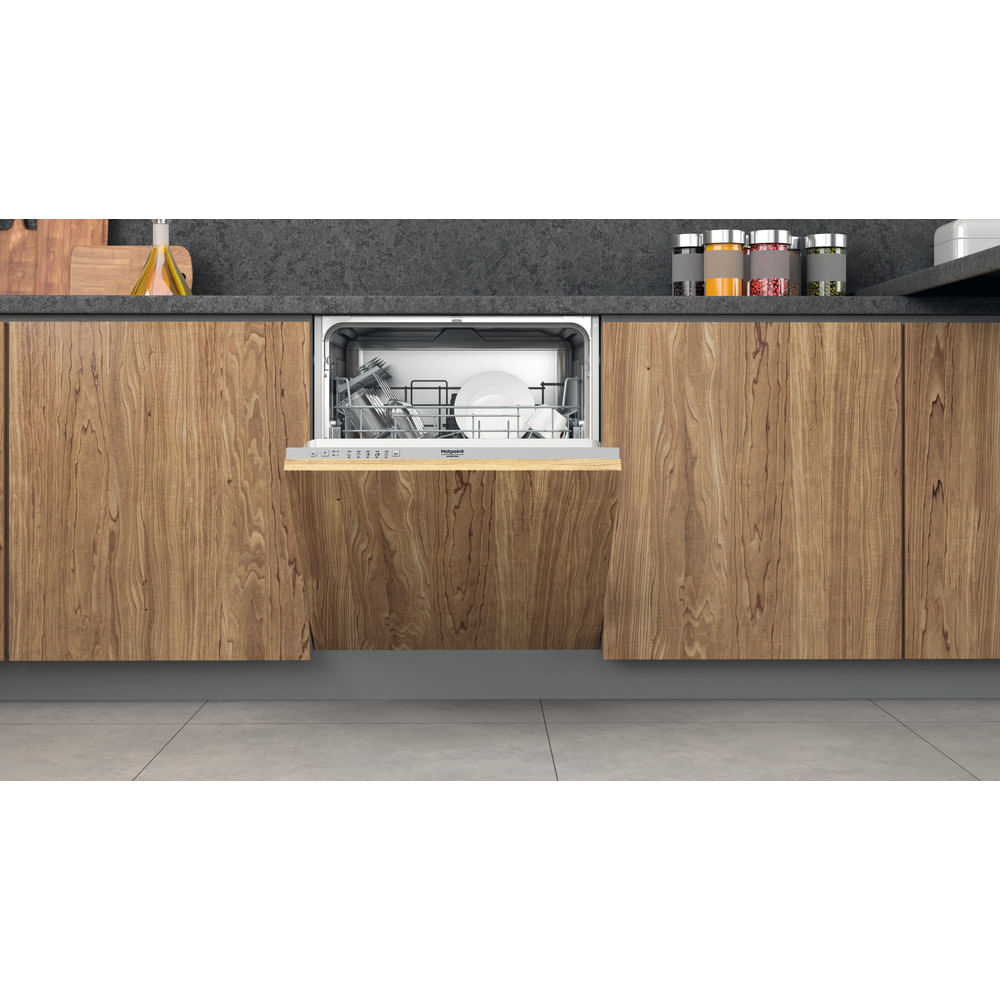 Un lavavajillas con la puerta abierta está integrado en armarios de madera. Dentro hay platos y cubiertos. Arriba hay especias y una tabla de madera.