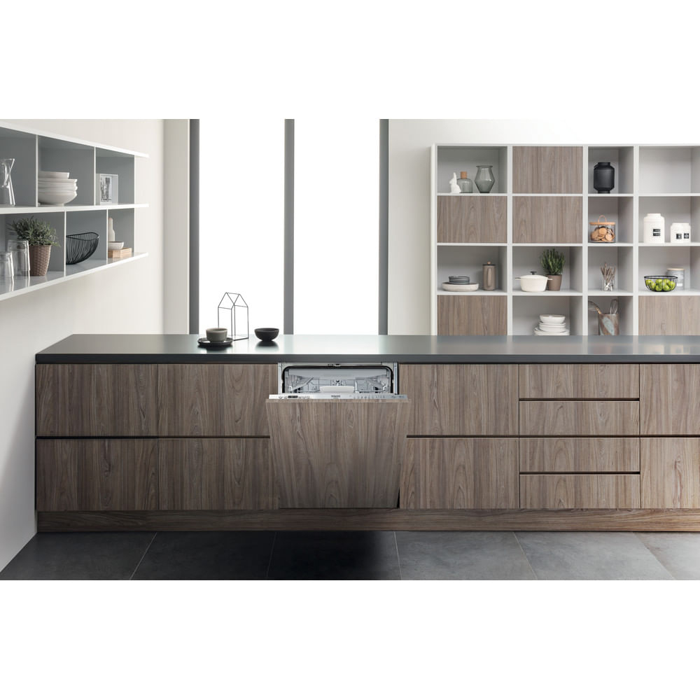 Cucina moderna con lavastoviglie aperta. Mobili e scaffali in legno con oggetti. Piano di lavoro e pavimento scuri. Finestra luminosa.