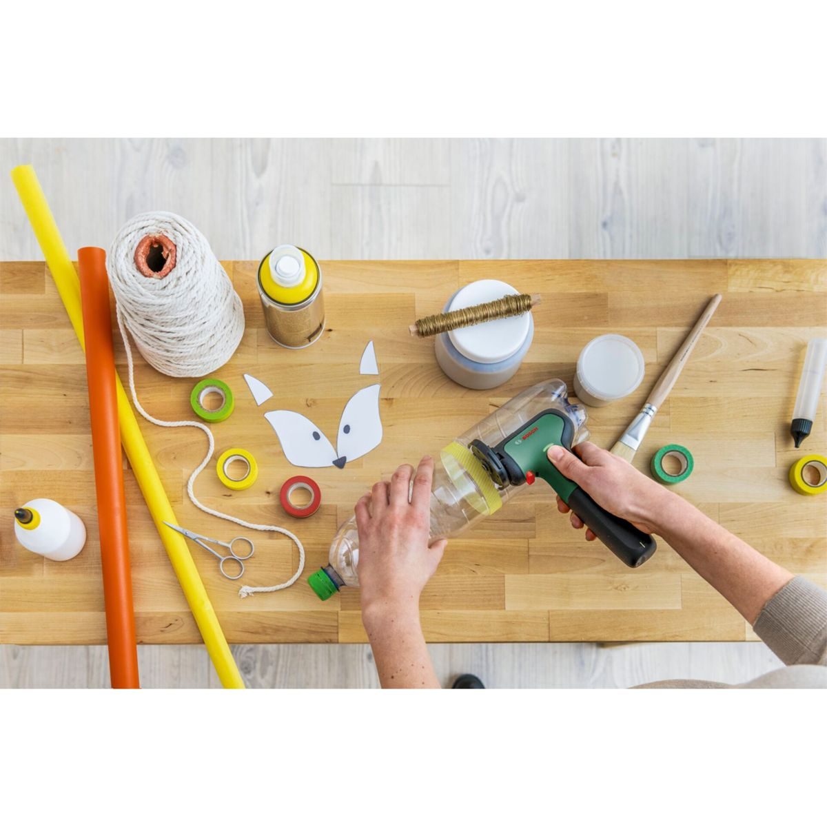 Des mains travaillent avec une bouteille en plastique et un outil sur une table en bois. Du matériel de bricolage tel que du ruban adhésif, des ciseaux et un renard en papier sont présents.