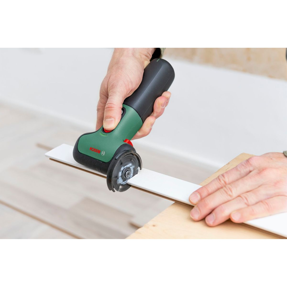 Une personne utilise une scie Bosch verte et noire pour couper une planche blanche. La lame coupe la planche sur une surface en bois.