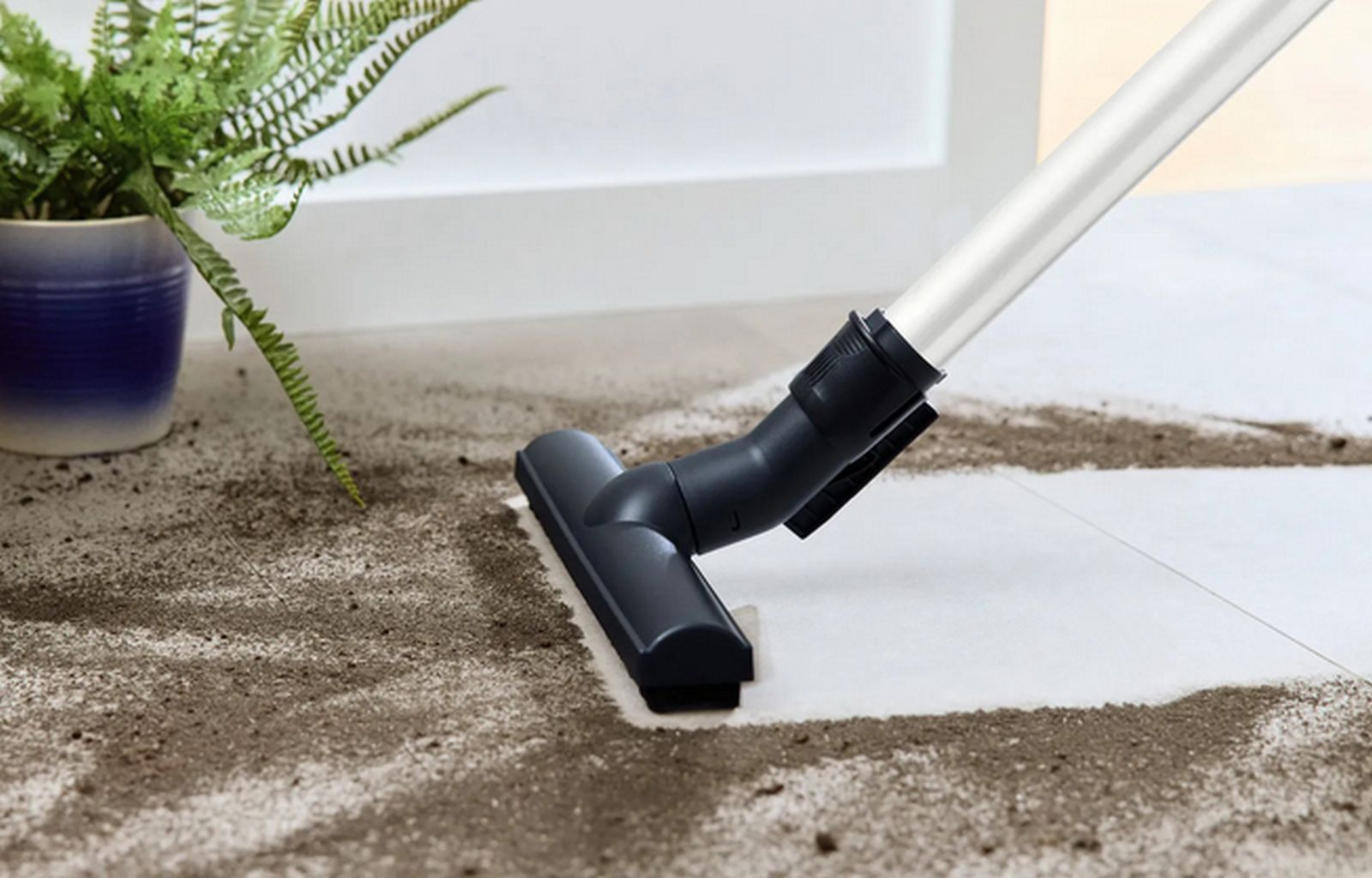 Odkurzacz czyści brud na białych płytkach. W tle roślina doniczkowa.