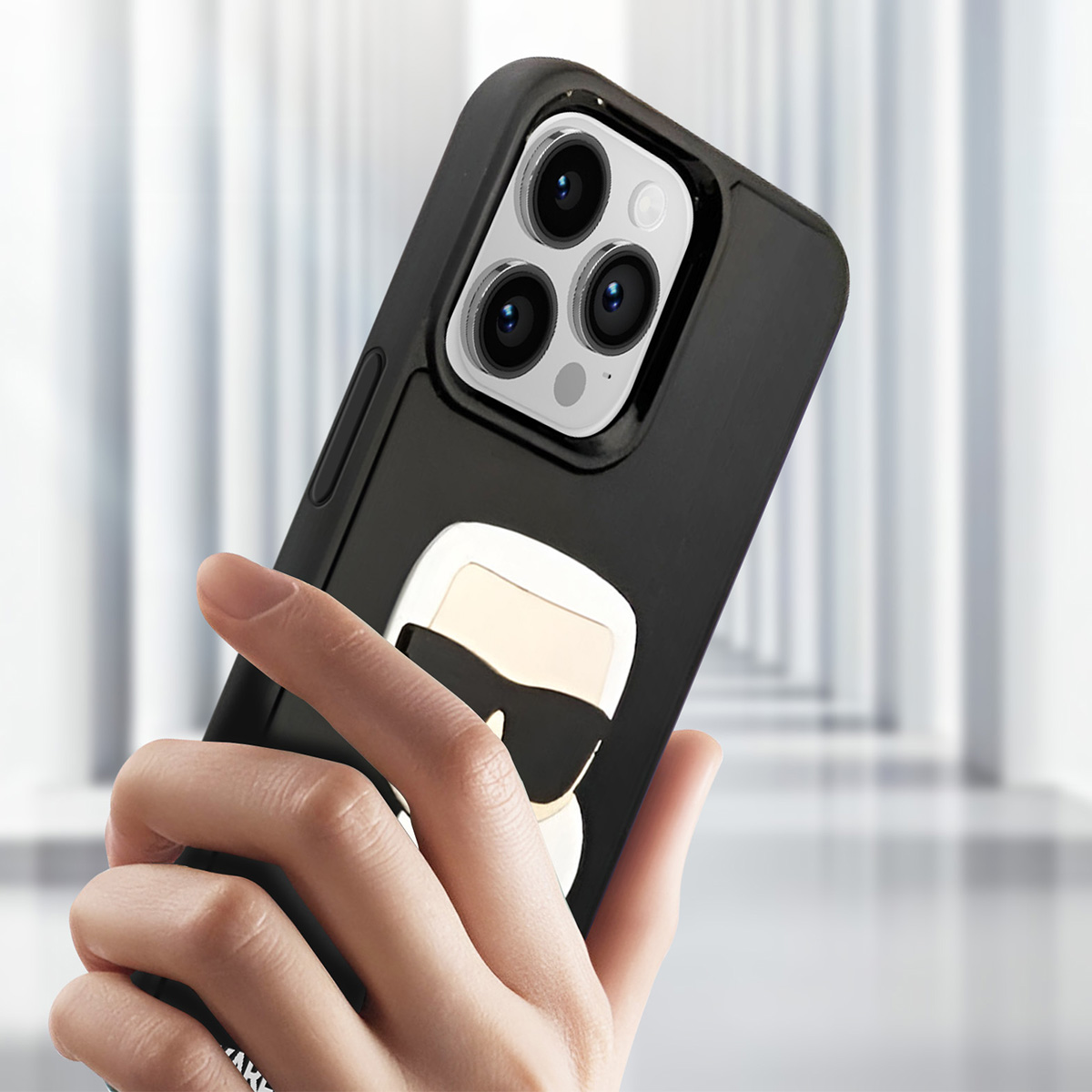 Una mano tiene una custodia nera per telefono con design Karl Lagerfeld. La custodia ha una fotocamera a tripla lente e sfondo sfocato.
