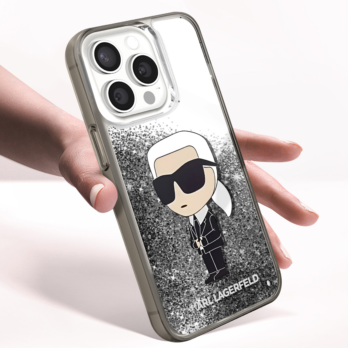Mano che tiene una custodia per telefono con un cartone animato di Karl Lagerfeld, con occhiali da sole e un abito nero, con sfondo glitter.