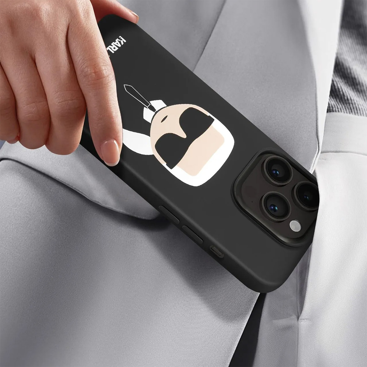 Una mano tiene un telefono nero con un design a cartoni animati, parzialmente in tasca. La custodia ha un design Karl Lagerfeld.