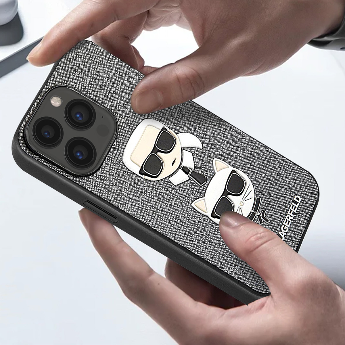 Mani che tengono una custodia per telefono con un design Karl Lagerfeld. La custodia è grigia con due personaggi dei cartoni animati. Il telefono ha tre lenti.