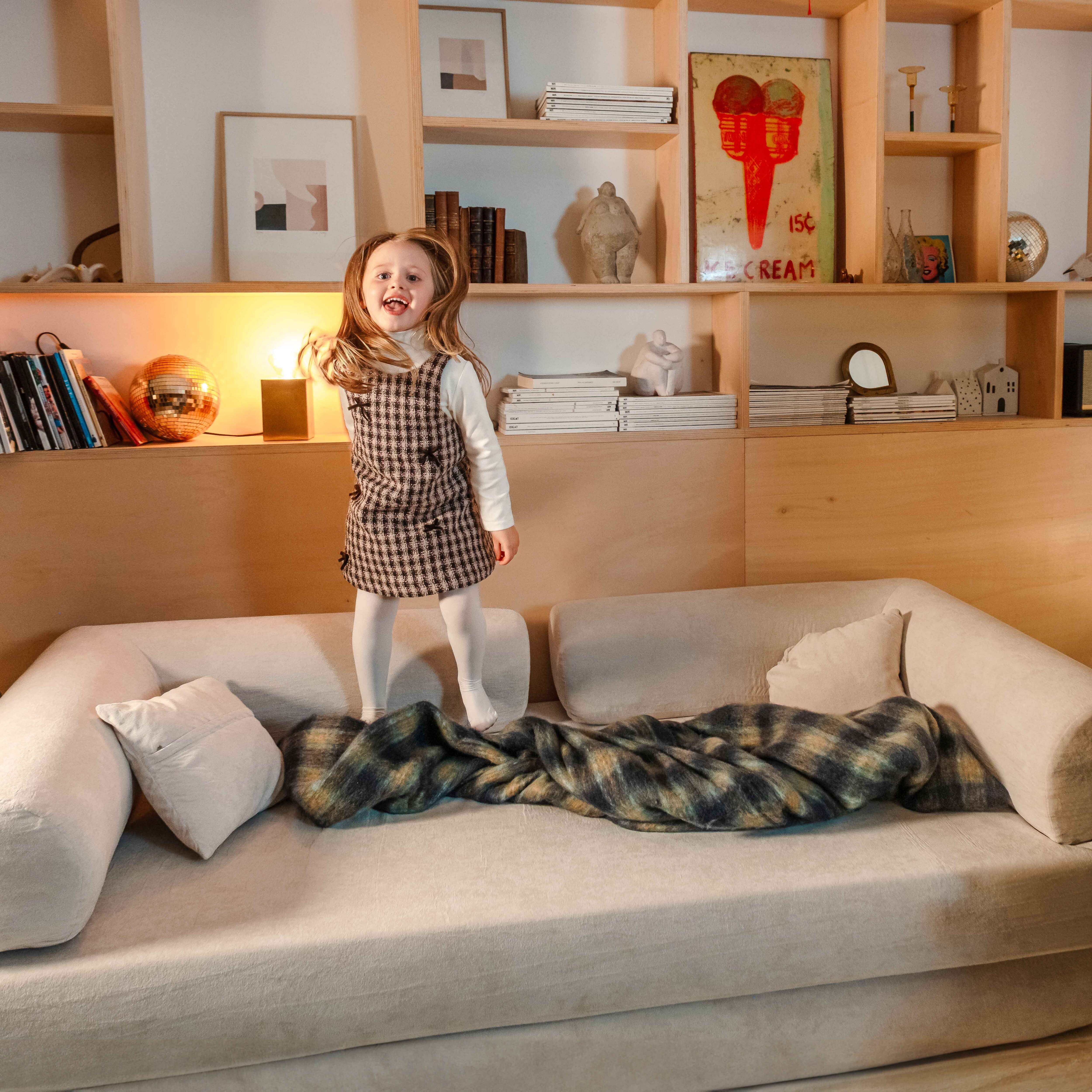 Ein Mädchen springt auf ein beiges Sofa, trägt ein Kleid. Eine karierte Decke und Kissen liegen auf dem Sofa. Regale im Hintergrund.