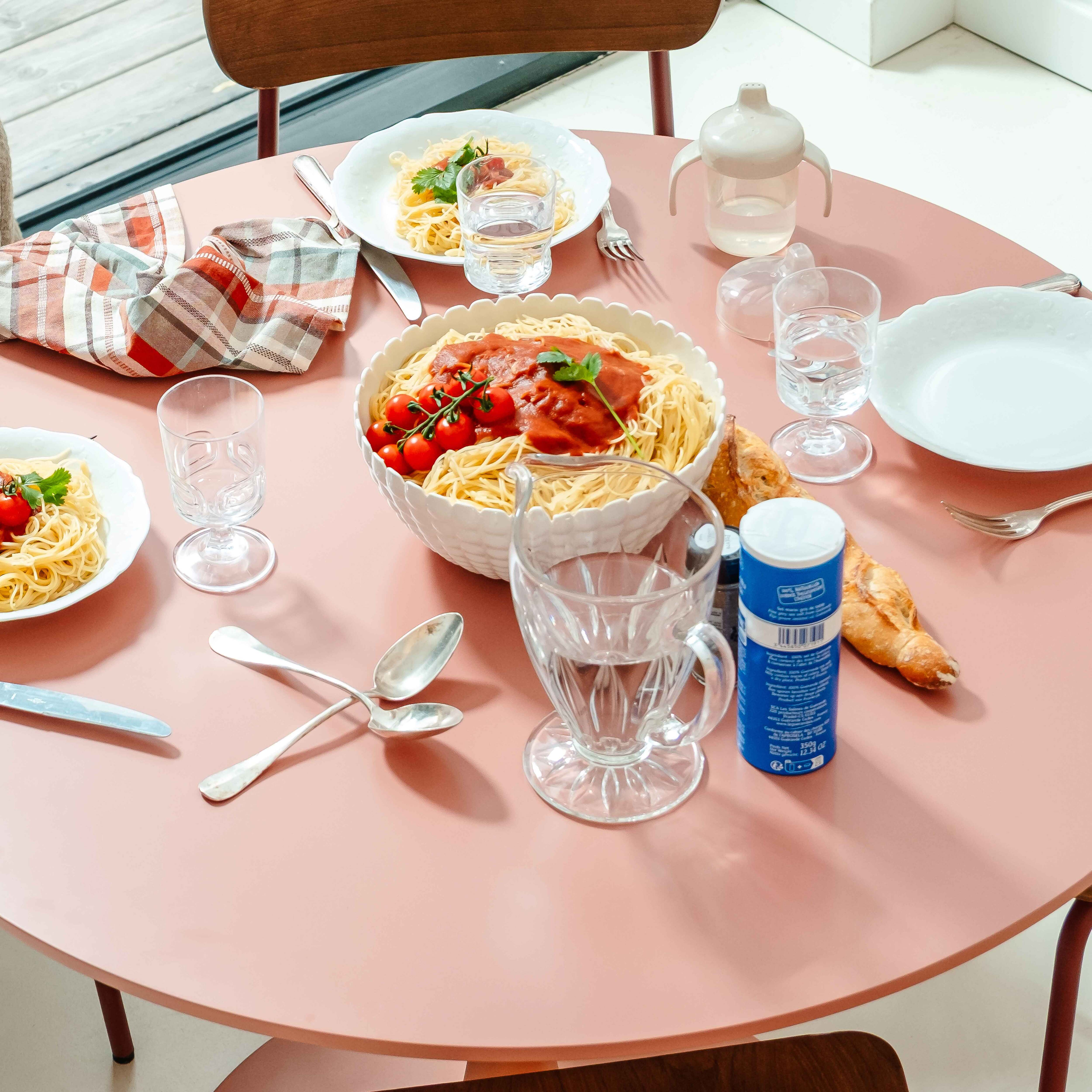 Ein runder Tisch mit Pasta, Tomaten, Wassergläsern und Utensilien. Eine Serviette und eine Babyflasche sind ebenfalls vorhanden.