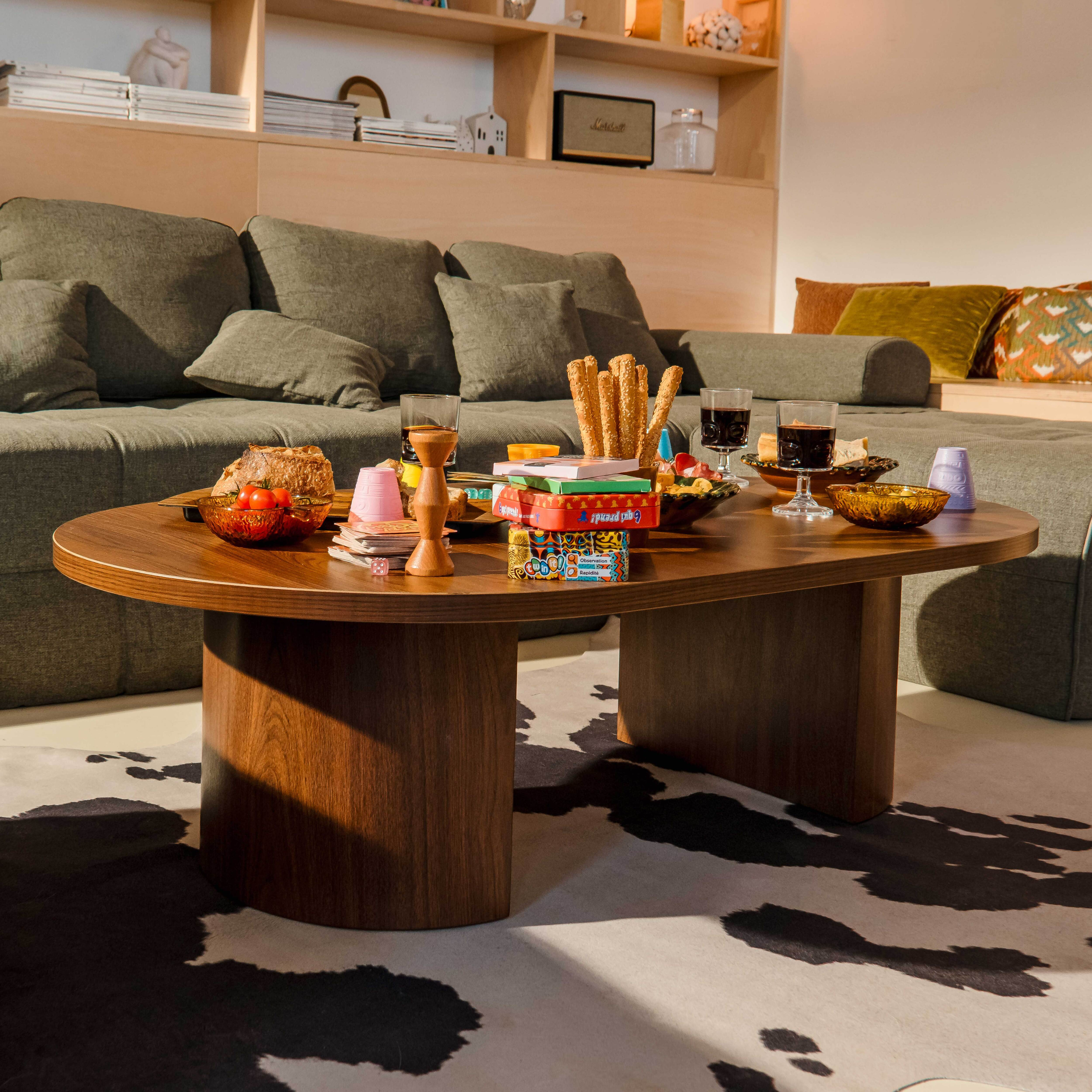 Ein Couchtisch mit Essen und Spielen steht vor einer grauen Couch. Ein Kuhfellteppich liegt darunter. Regale sind hinten.