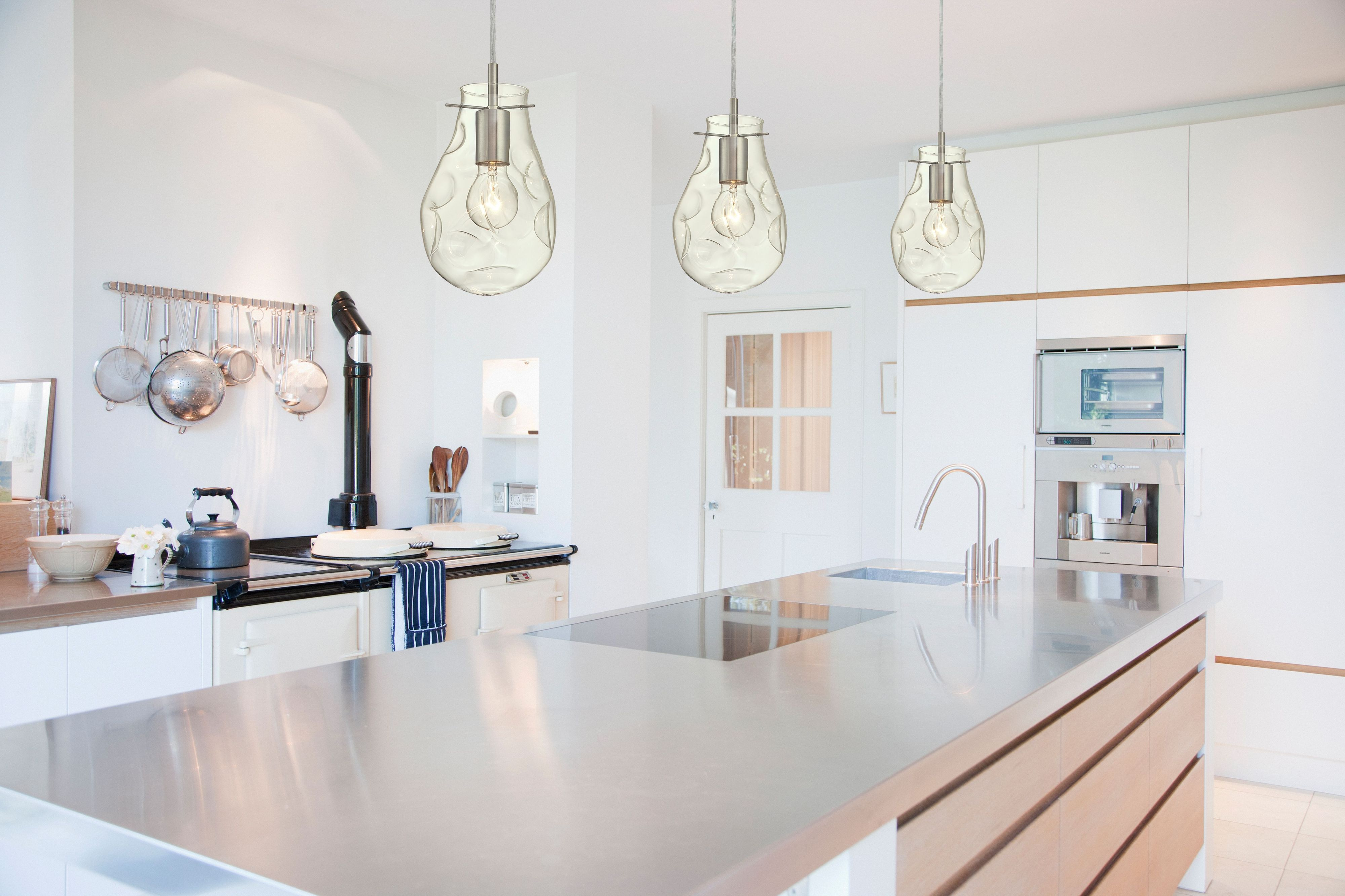 Moderne Küche mit Edelstahlinsel, Hängeleuchten, Herd und weißen Schränken. Fokus auf Design und Funktionalität.