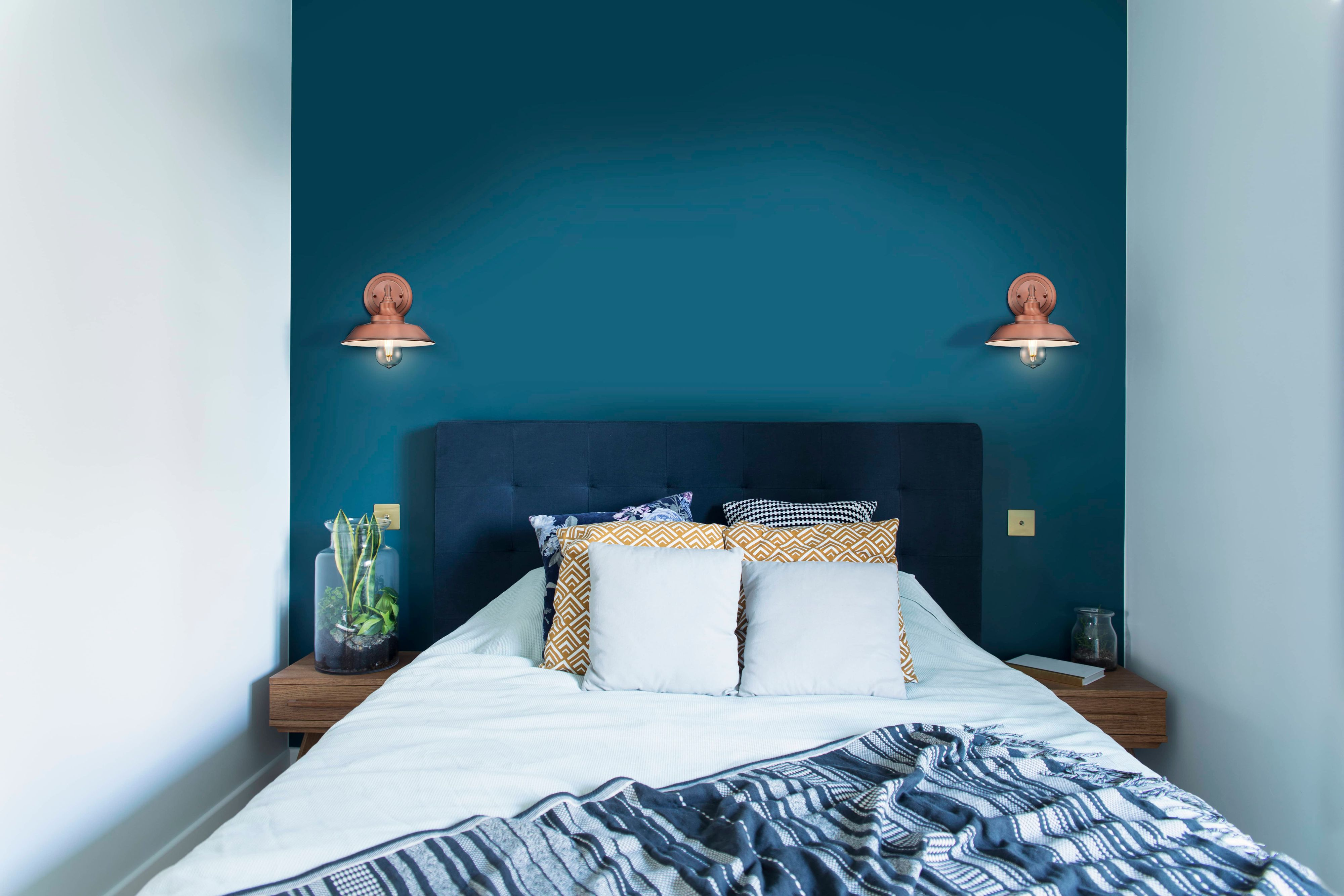 Schlafzimmer mit blauer Wand, Bett mit Kissen, zwei Lampen und Pflanzen. Eine Decke liegt auf dem Bett.