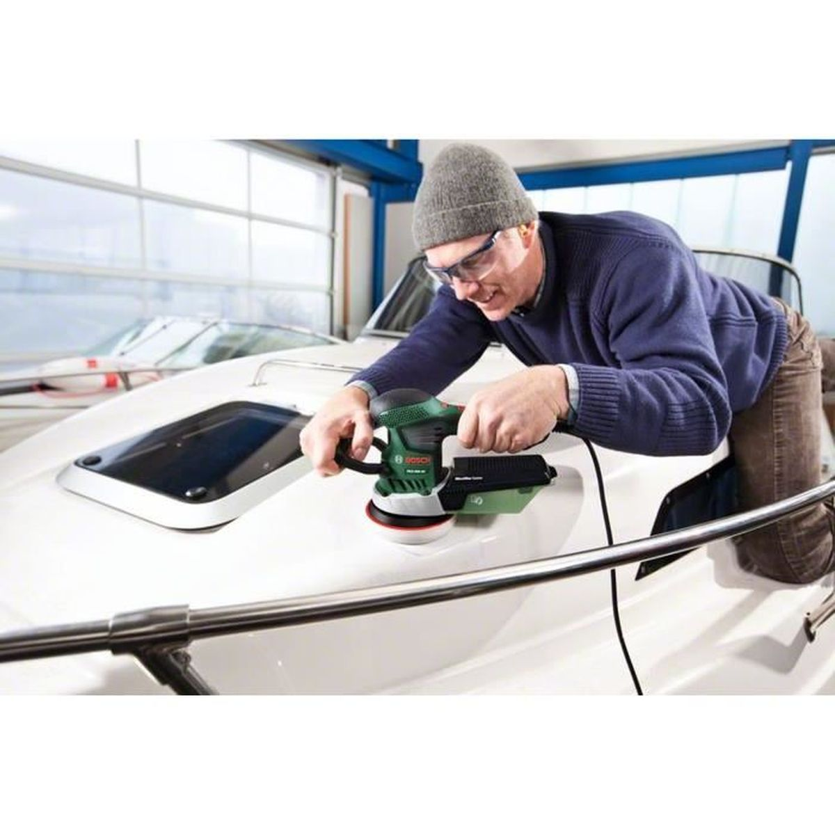 Un homme polit un bateau blanc avec une ponceuse électrique. Il porte un bonnet, des lunettes et un pull bleu. L'arrière-plan est un hangar à bateaux.