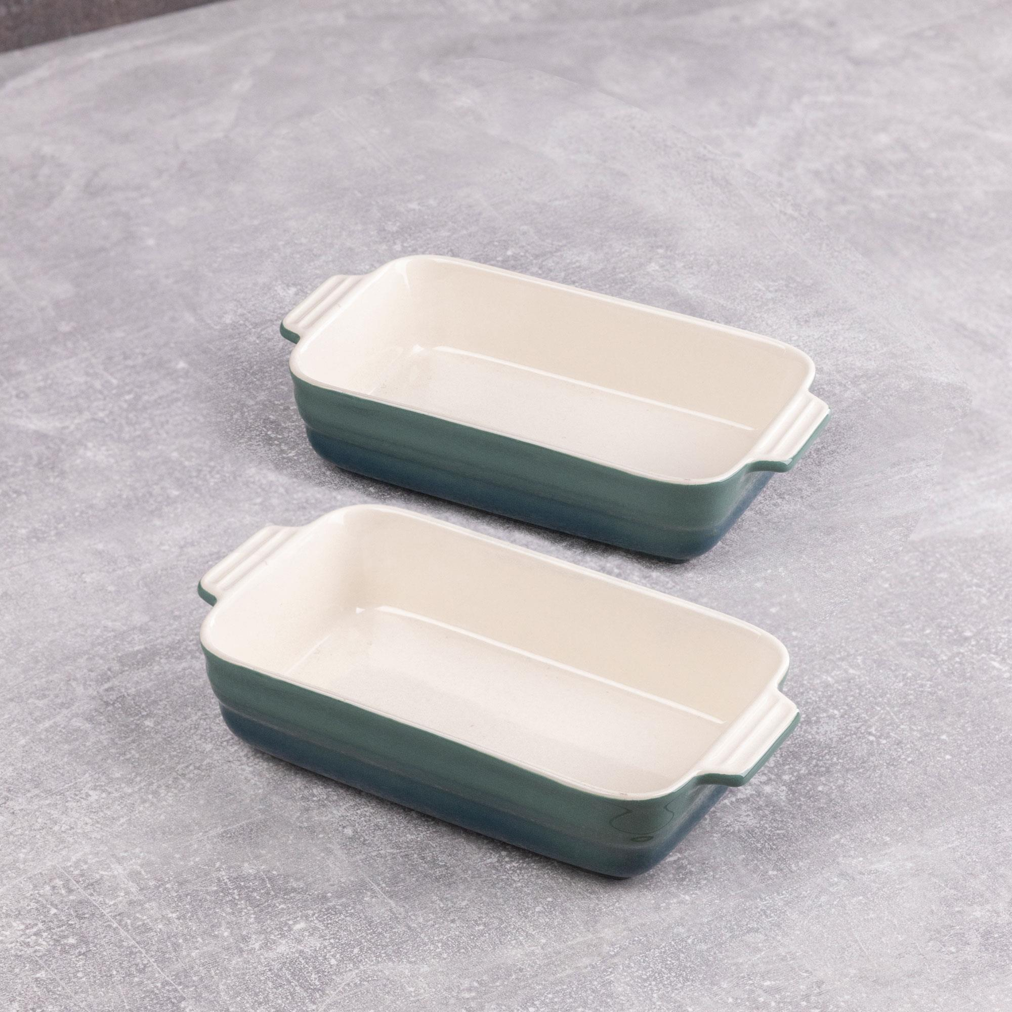 Dos fuentes rectangulares para hornear con asas sobre una superficie gris texturizada. Los platos son verde azulado y crema.