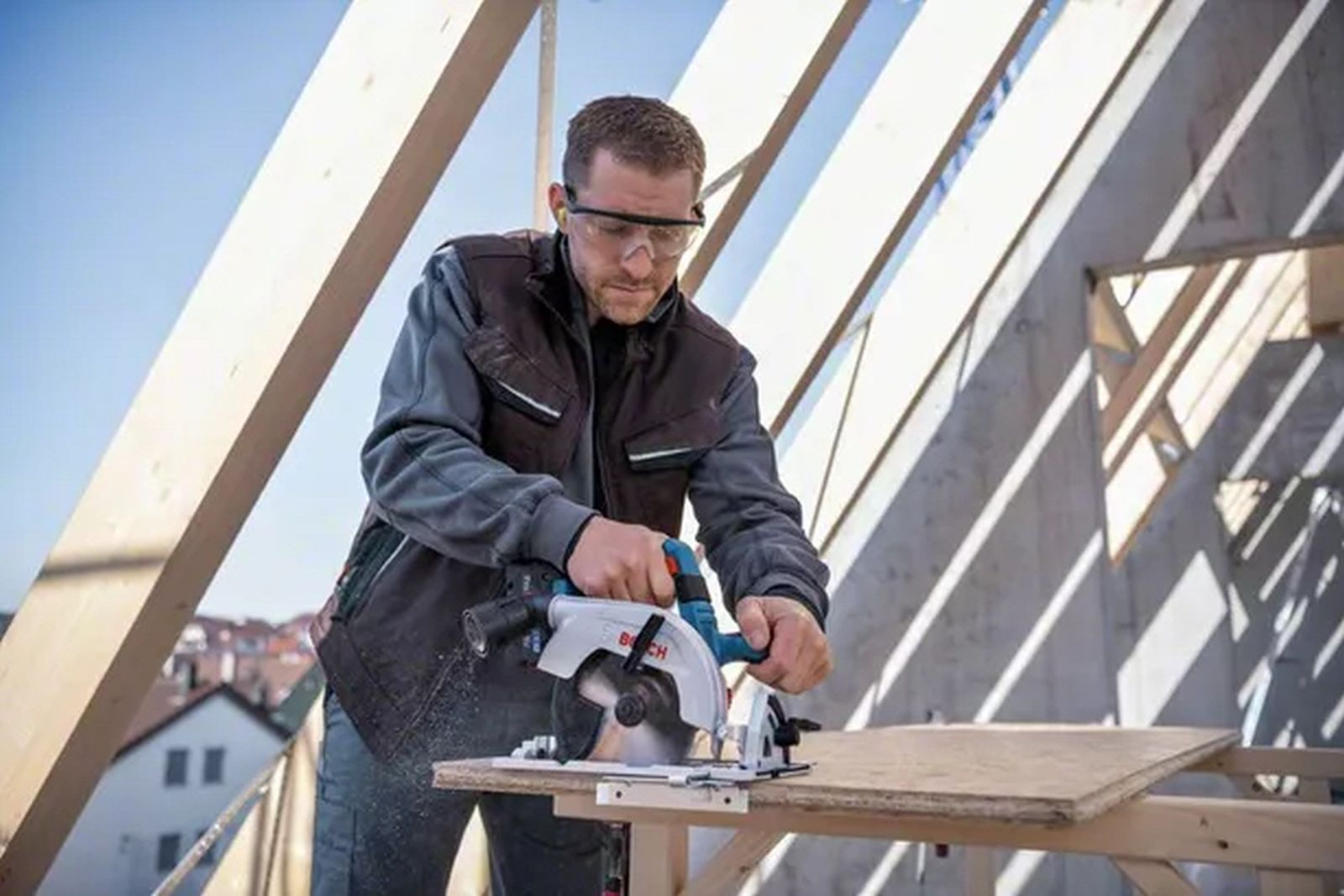 Mężczyzna używa piły tarczowej do cięcia drewna. Ma na sobie okulary ochronne. Scena rozgrywa się w częściowo zbudowanej konstrukcji.