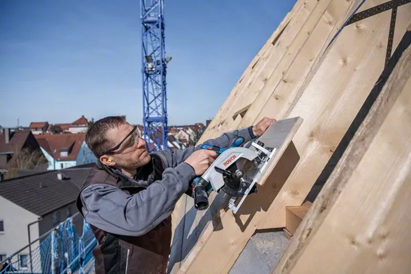 Mężczyzna używa piły tarczowej na drewnianej konstrukcji dachu. Niebieski dźwig w tle. Domy widoczne w oddali.