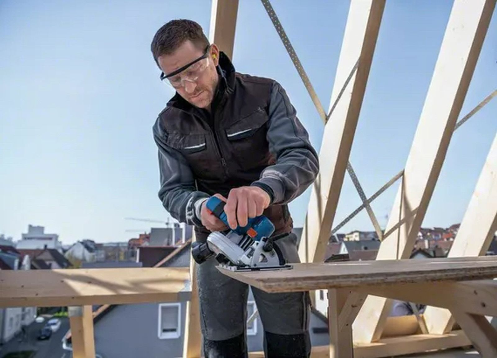 Mężczyzna w sprzęcie ochronnym używa piły tarczowej do drewna. Budynki i błękitne niebo w tle.