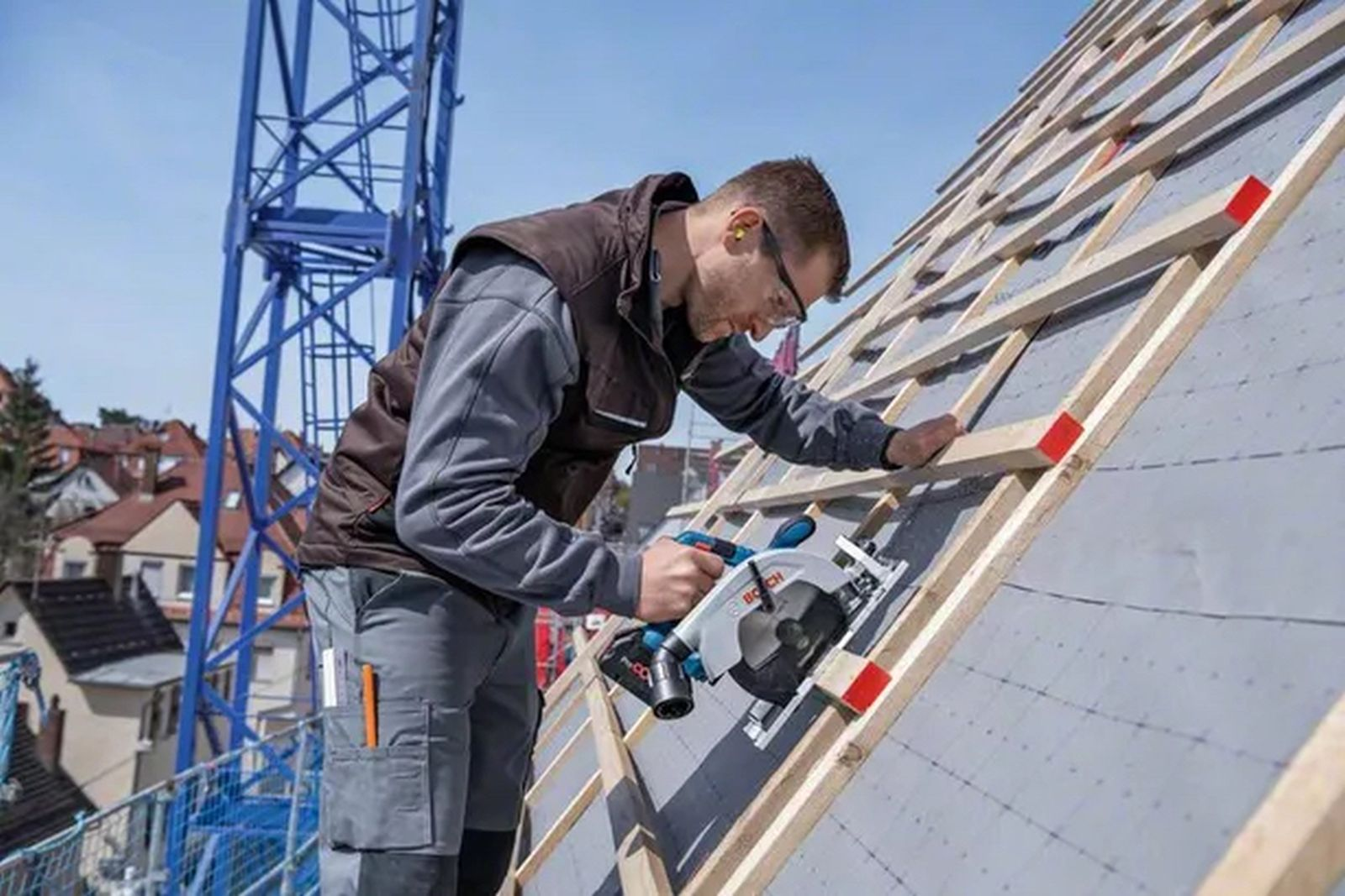 Mężczyzna używa piły tarczowej na dachu. Żuraw i domy w tle. Niebo jest niebieskie.
