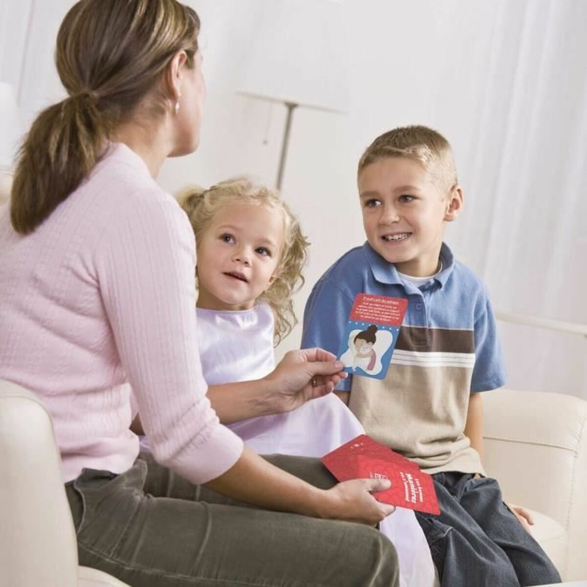 Una donna mostra delle carte a due bambini su un divano. I bambini guardano le carte. Lo sfondo ha una lampada e una tenda bianca.