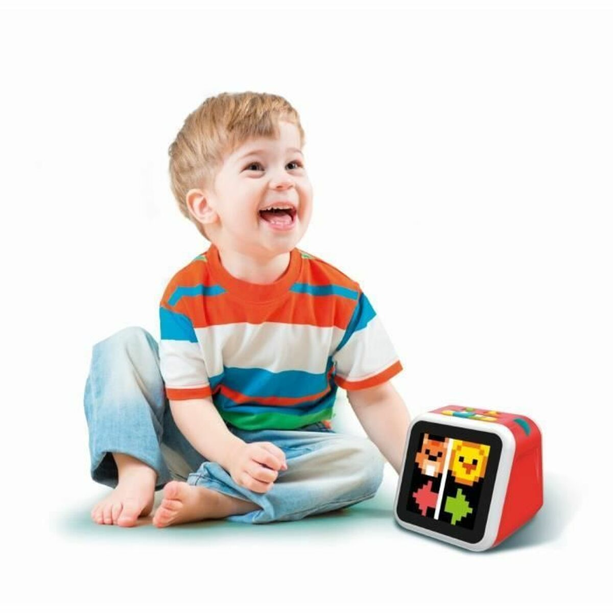 Un bambino sorridente con una maglietta a righe è seduto accanto a un cubo rosso con facce di animali pixelate sullo schermo.