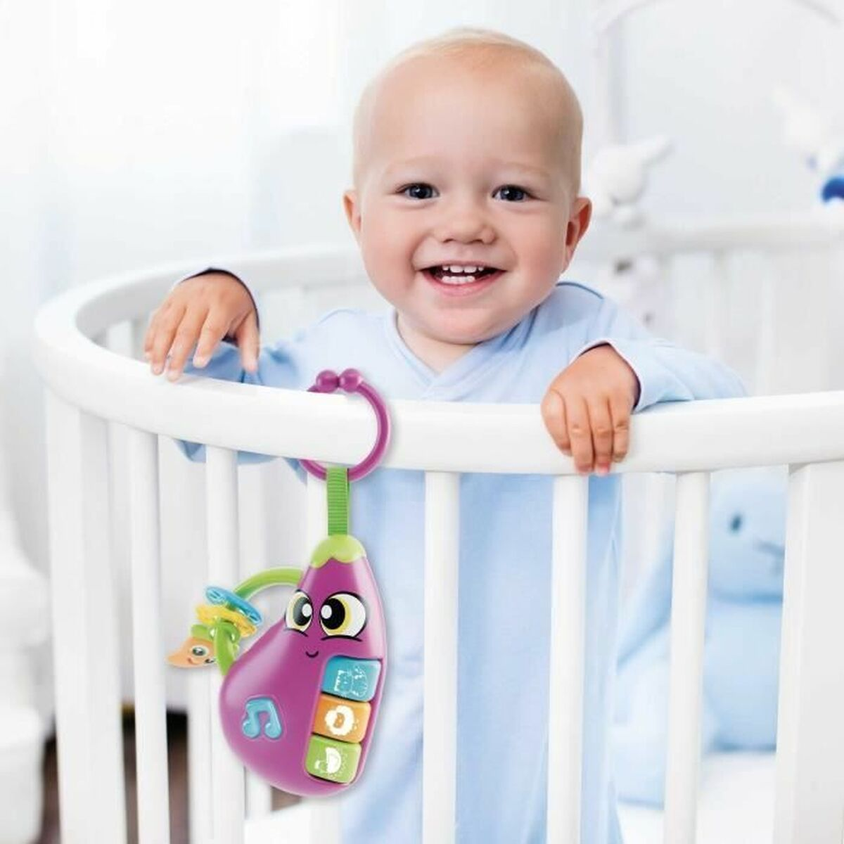 Ein lächelndes Baby in einem Kinderbett mit einem Spielzeug-Aubergine. Das Spielzeug ist lila, grün und gelb. Das Baby trägt blau.