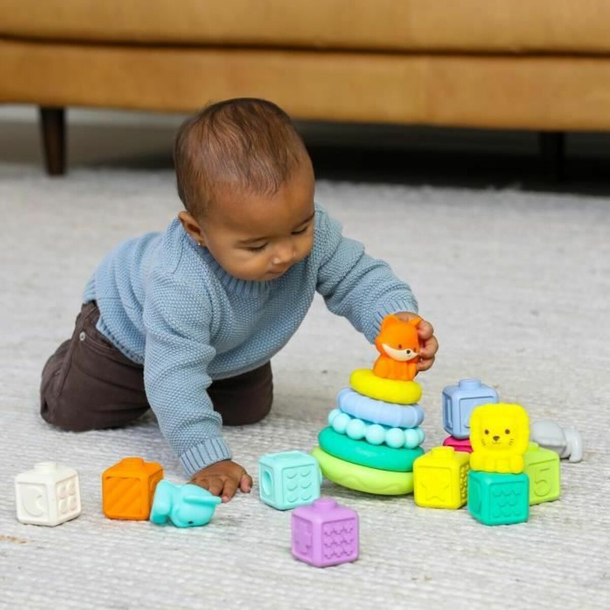 Un bébé joue avec des anneaux empilables et des blocs colorés. Le bébé porte un pull bleu et un pantalon marron, sur un tapis blanc.