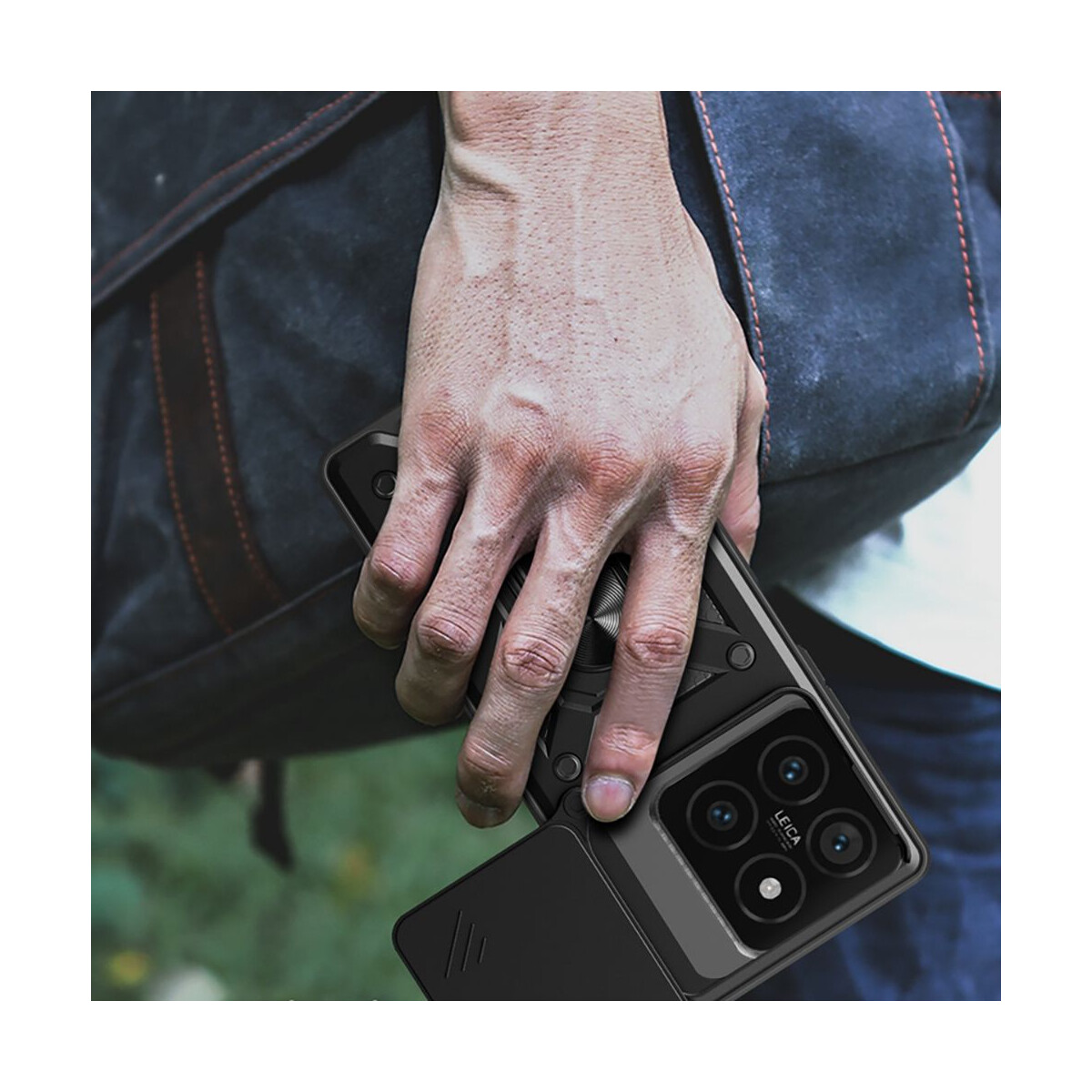 Eine Hand hält ein schwarzes Telefon mit geöffneter Kameraabdeckung. Im Hintergrund eine dunkle Tasche. Die Handyhülle hat einen Ringgriff.