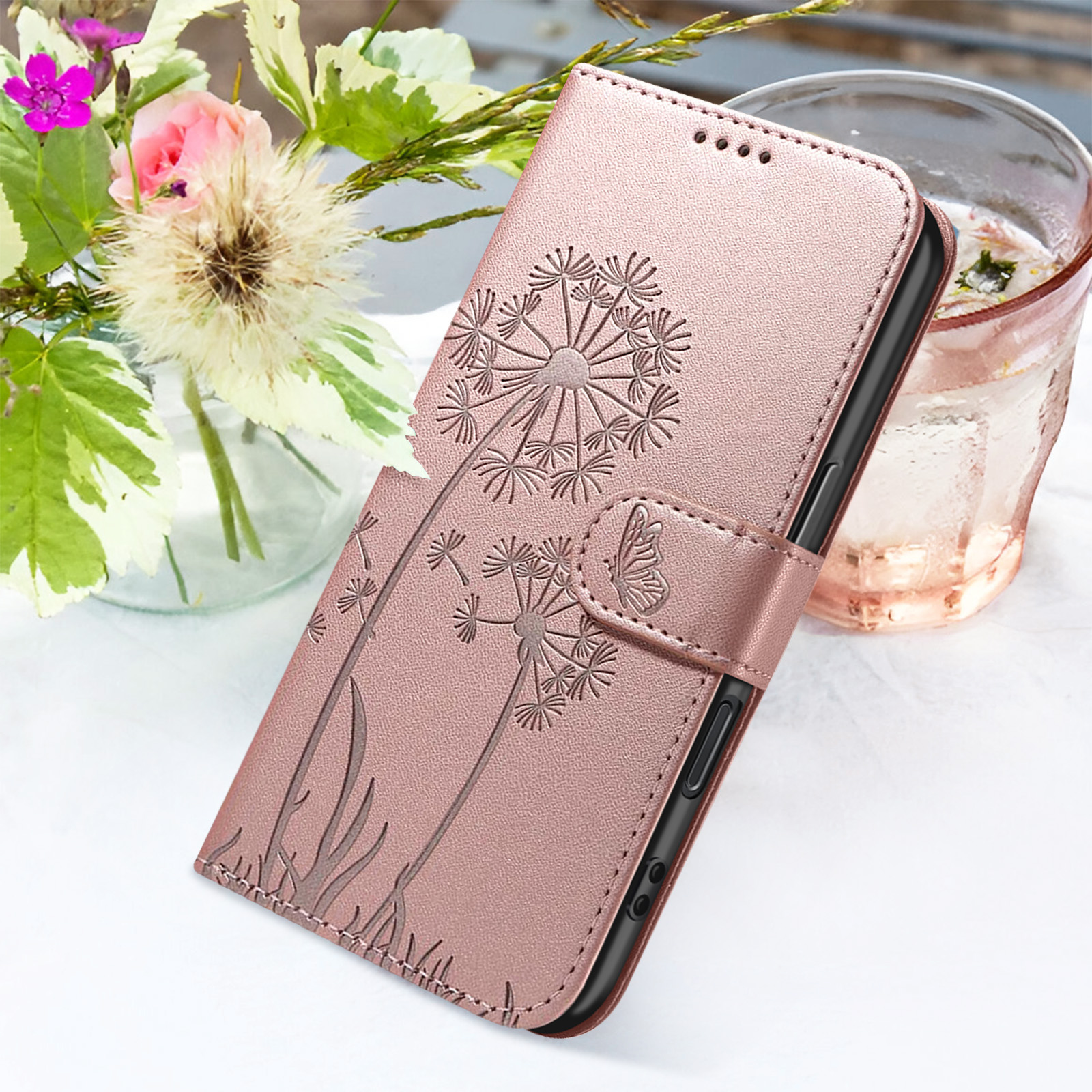 Eine rosa Handyhülle mit Pusteblumen-Design, ein Glas Wasser und Blumen in einer Vase auf weißer Oberfläche.