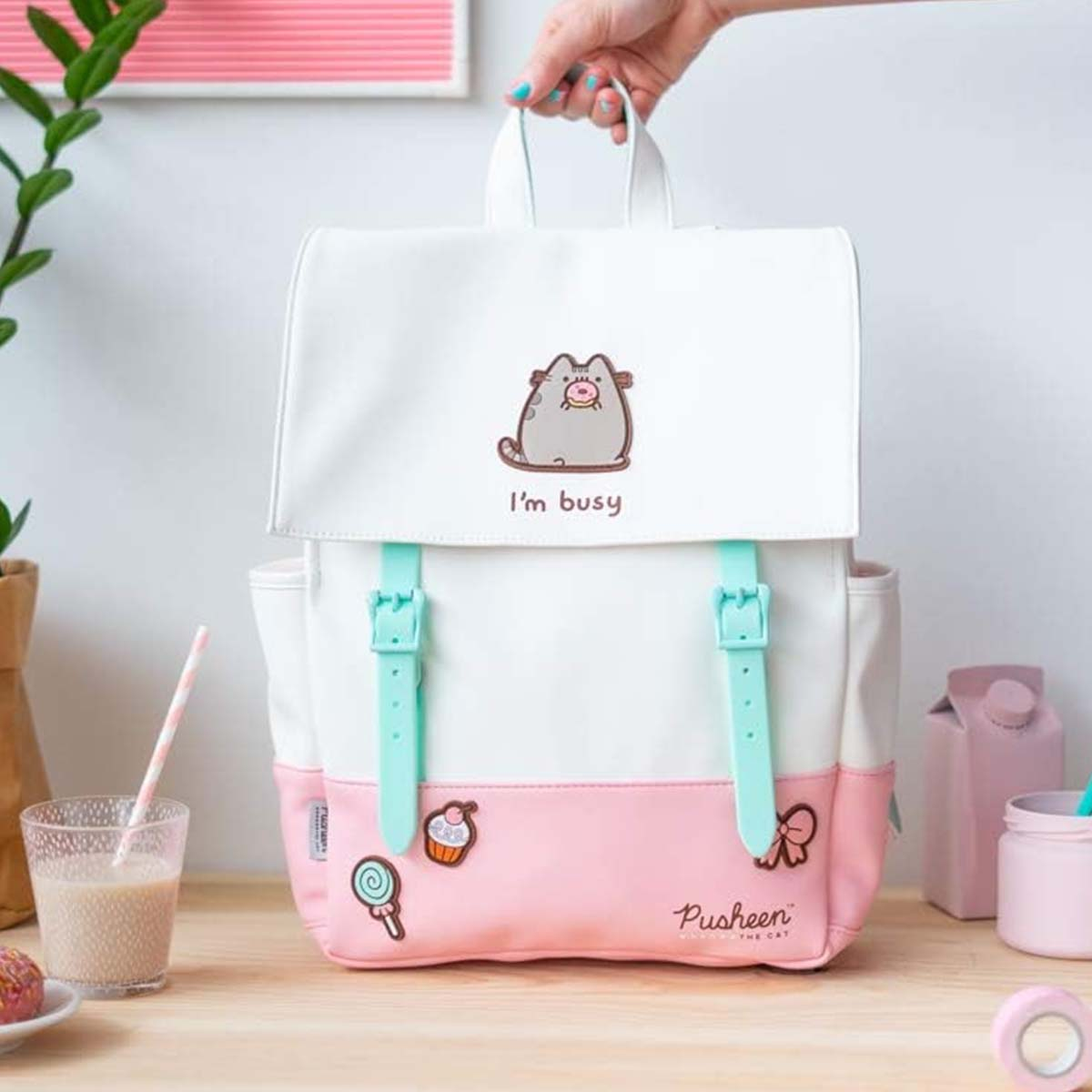 Zaino bianco con base rosa, grafica del gatto Pusheen e cinghie blu. Una mano tiene la maniglia. Bevande e decorazioni sullo sfondo.