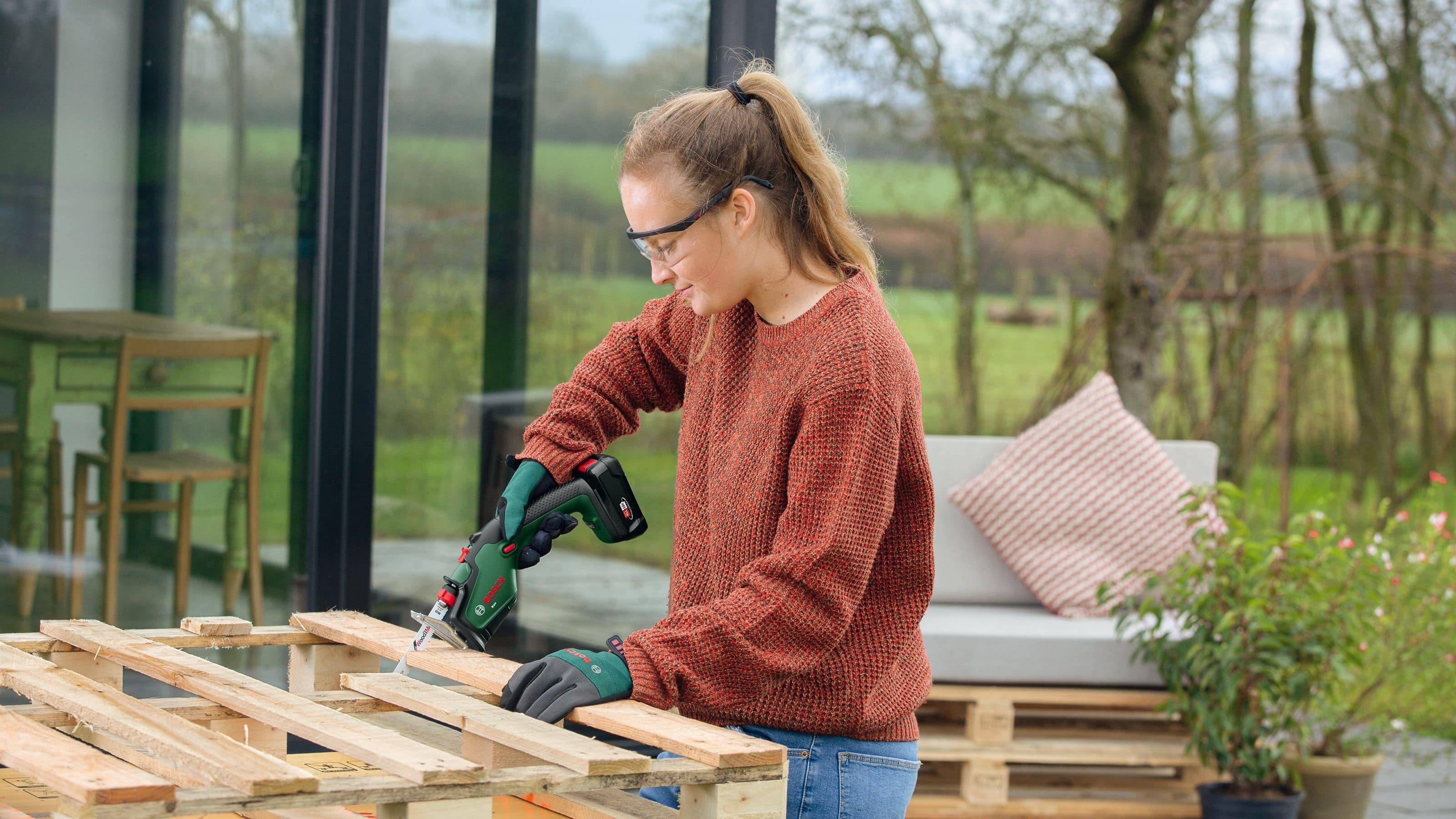 Eine Frau sägt im Freien Holz auf einer Palette. Sie trägt eine Schutzbrille, einen braunen Pullover und Handschuhe. Hintergrund: Bäume und eine Terrasse.