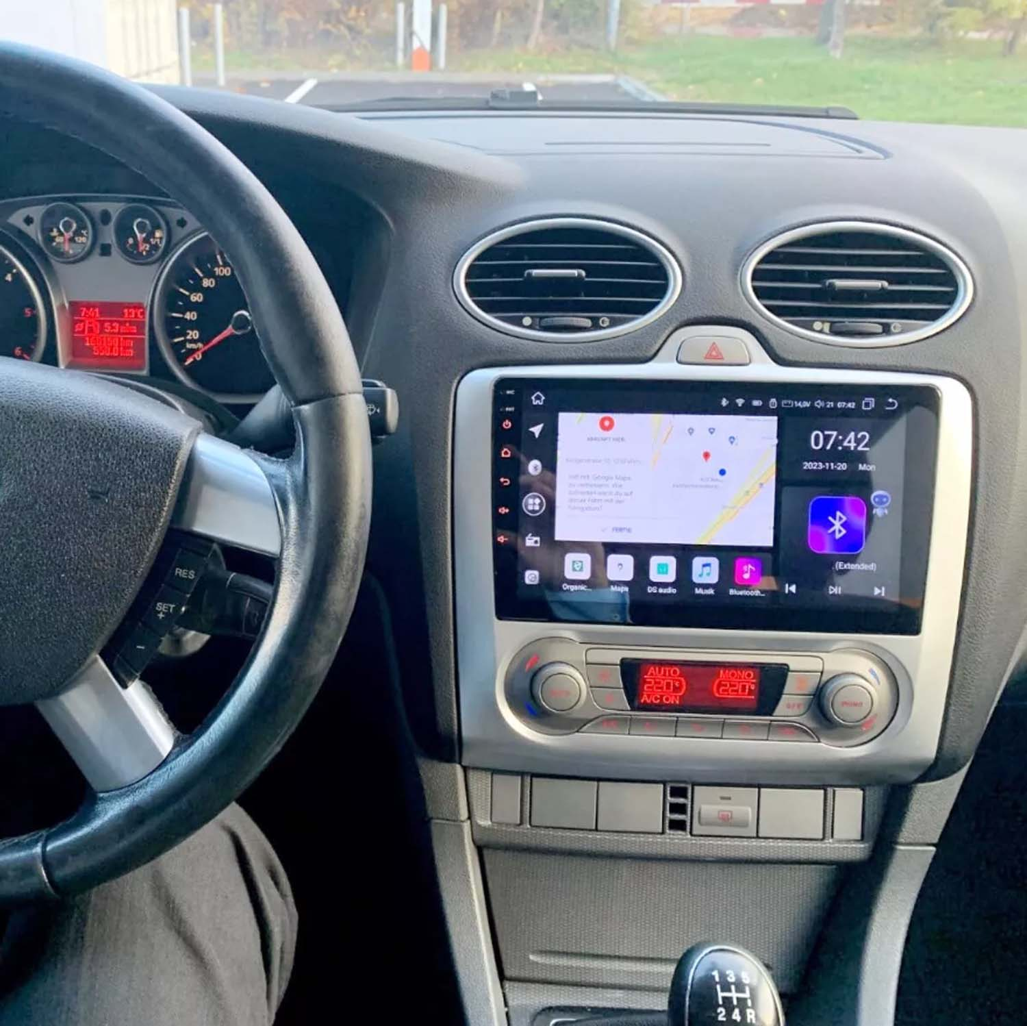 Vista interna dell'auto. Volante, cruscotto con display digitale che mostra mappa e ora, cambio visibile.