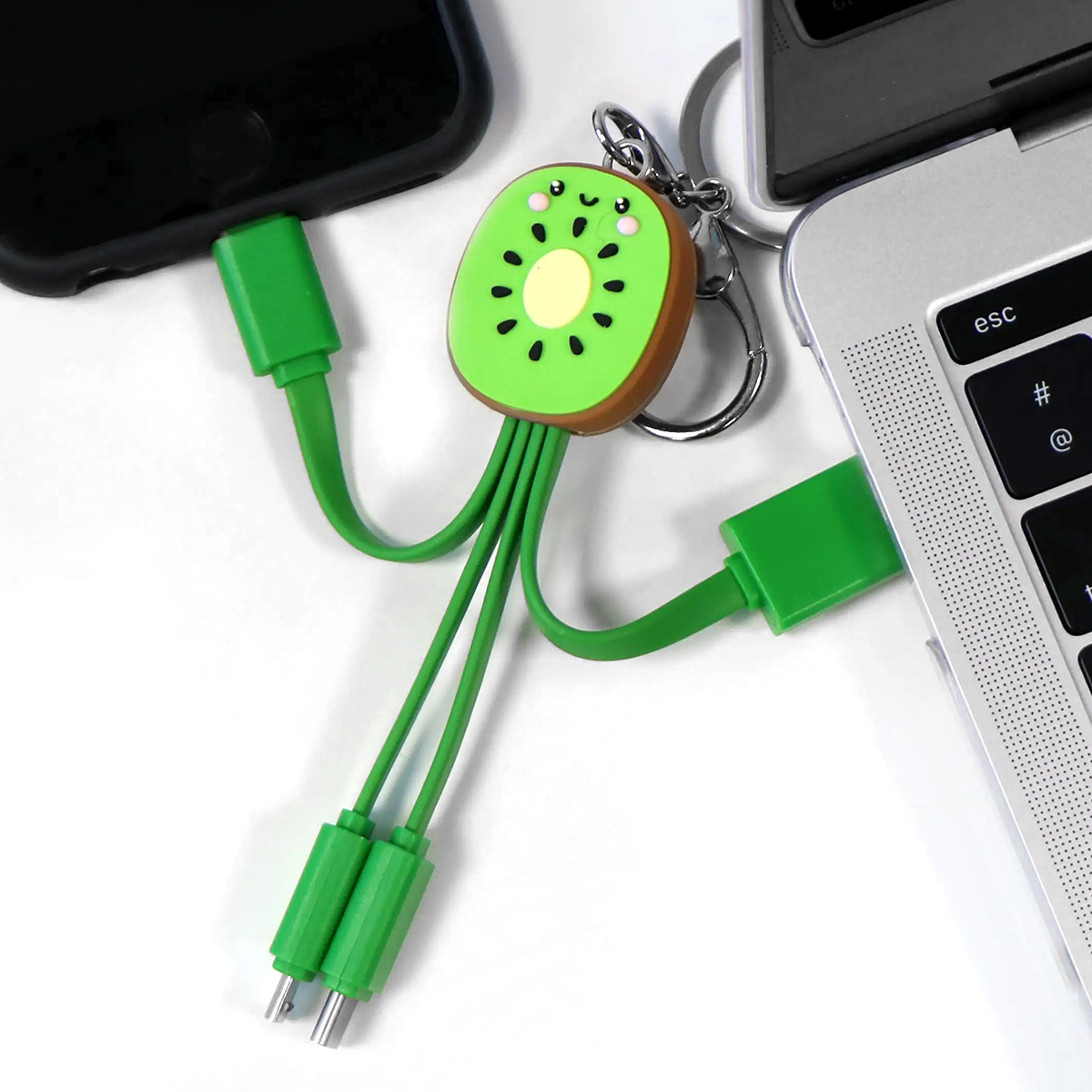 Un cavo di ricarica a forma di kiwi è collegato a un telefono e a un laptop. Il cavo è verde con più connettori. Superficie bianca.