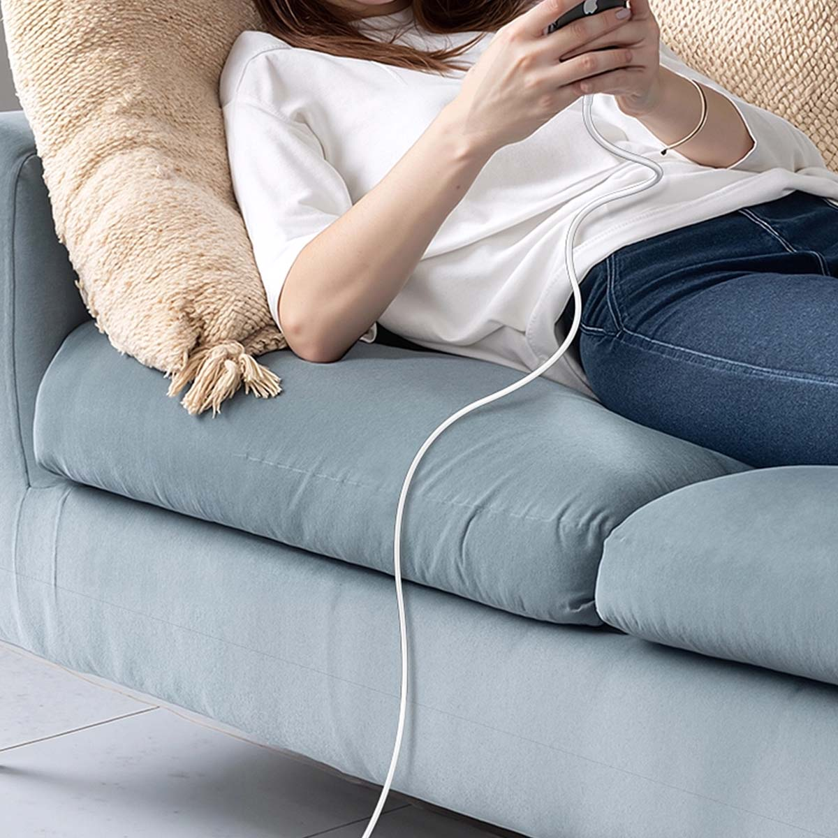 Osoba na niebieskiej kanapie używa telefonu. Ma na sobie białą koszulę i dżinsy. Za nią jest beżowa poduszka. Widoczny jest biały kabel.