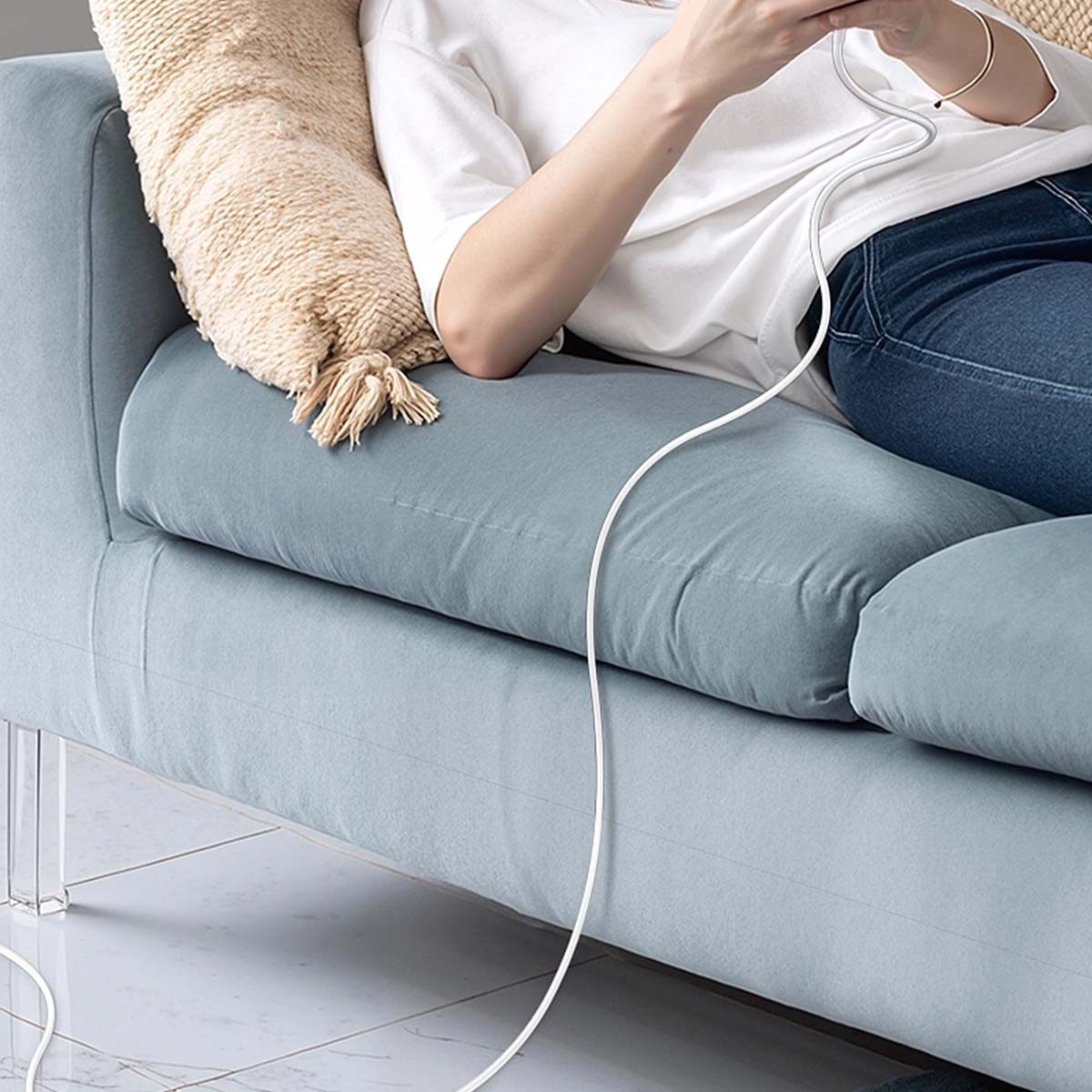 Osoba na niebieskiej kanapie korzysta z telefonu. Beżowa poduszka jest po lewej stronie. Biały przewód jest podłączony.