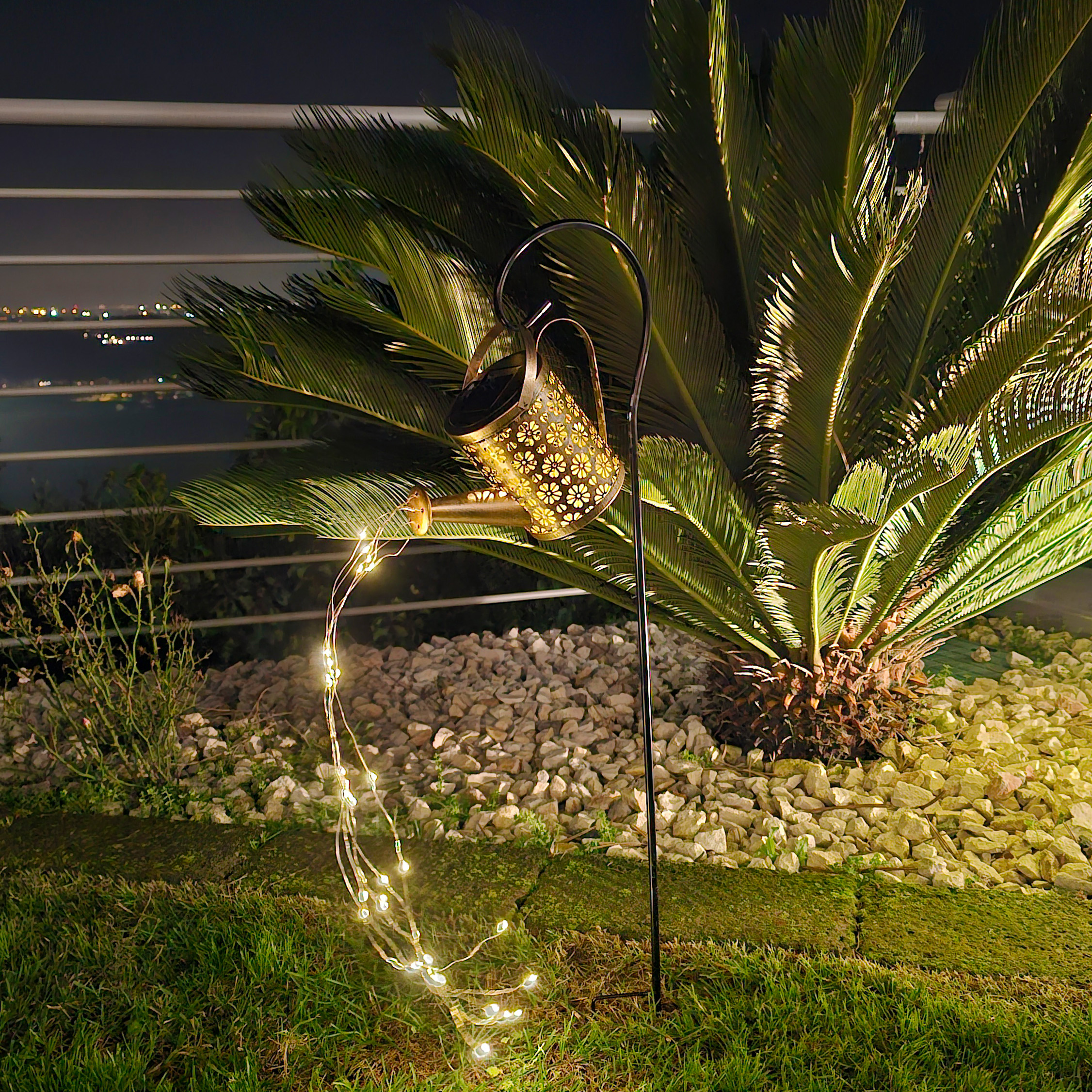 Una lampada decorativa a forma di annaffiatoio in un giardino di notte. Luci fatate e palme sono presenti nella scena.