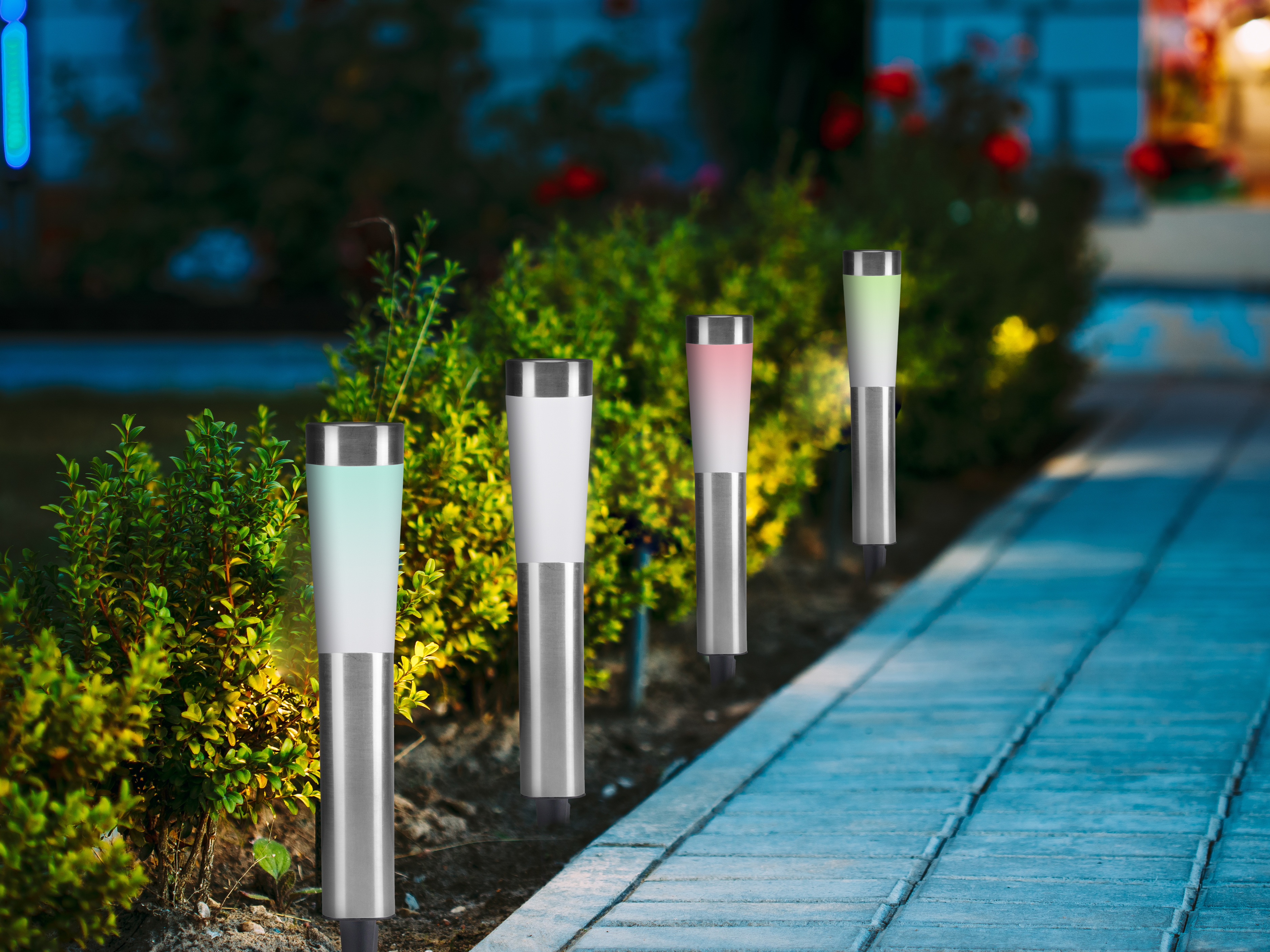 Quattro luci da giardino illuminate con cime di diversi colori, ambientate in un giardino con piante e un sentiero di pietra blu.