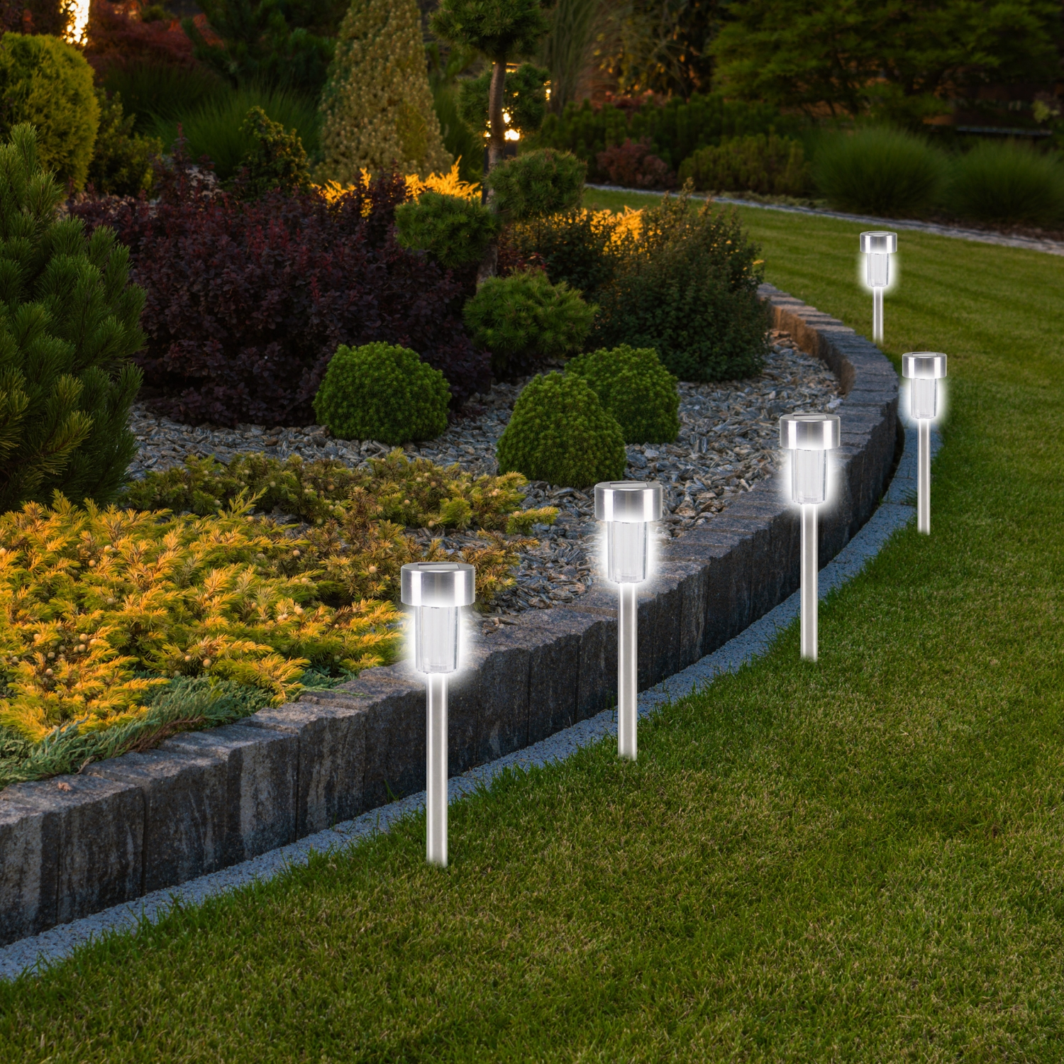 Scena del giardino al crepuscolo. Luci del sentiero illuminate davanti a un muro di pietra e vari cespugli. Erba verde lussureggiante.
