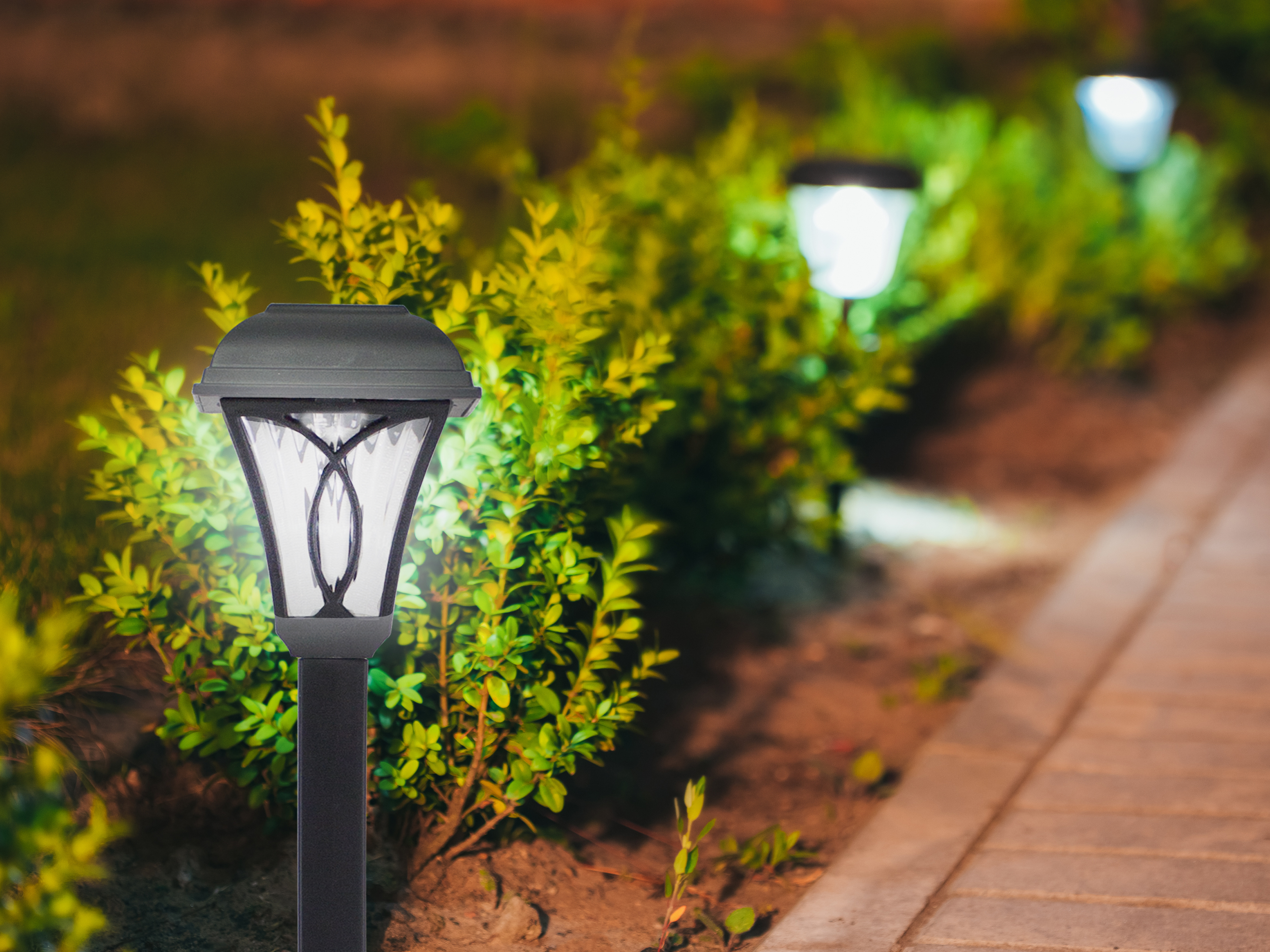 Una lampada da giardino illuminata a fuoco. Altre lampade sullo sfondo. Arbusti verdi e un sentiero in mattoni.