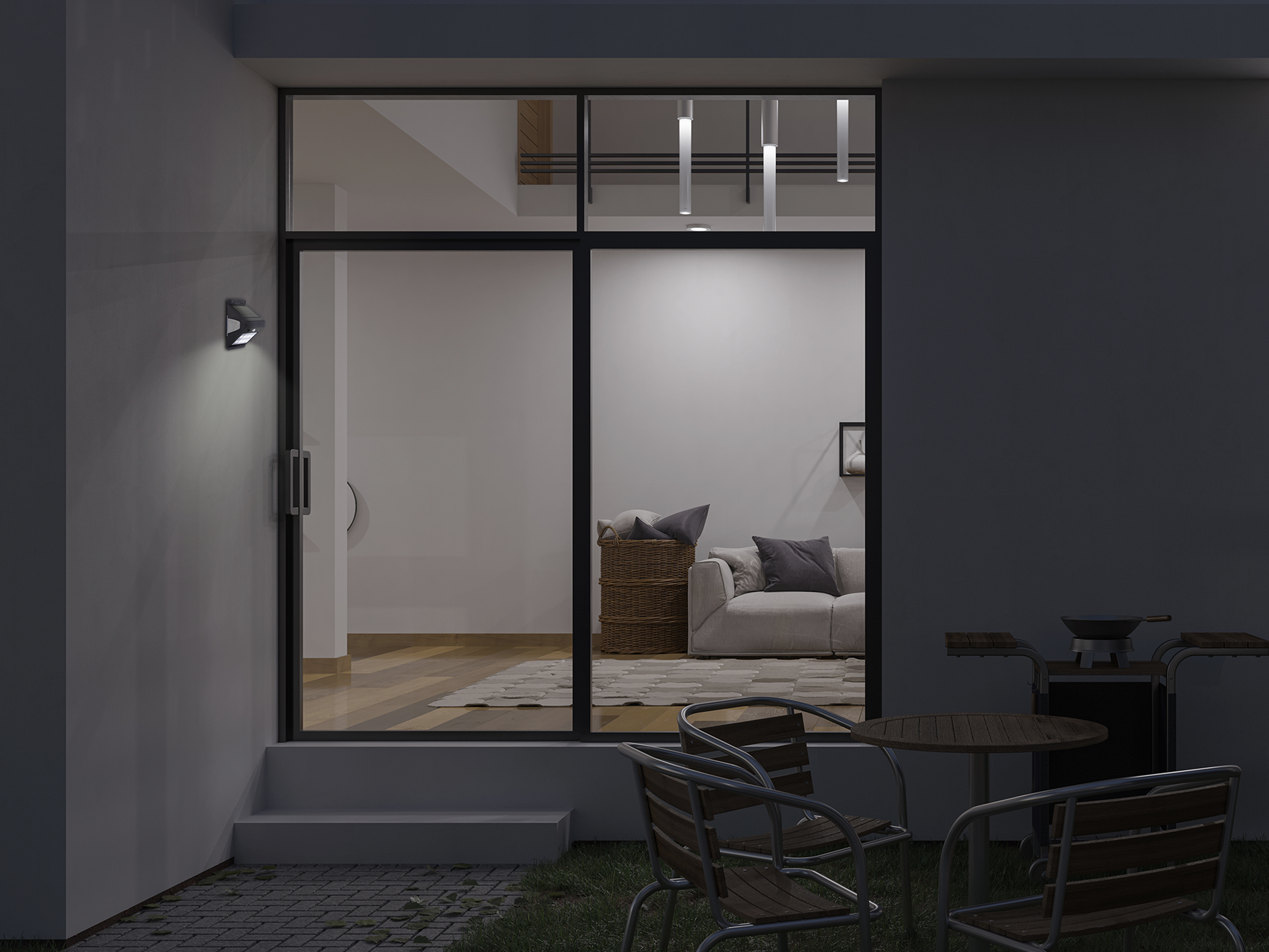 Vista notturna di un patio con mobili, barbecue e vista su un soggiorno illuminato. Una lampada da parete è accesa.