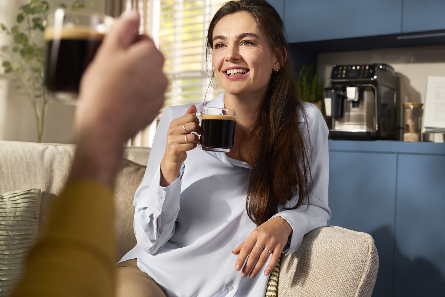 Kobieta uśmiecha się, trzymając kawę. Inna osoba trzyma kawę na pierwszym planie. Ekspres do kawy i niebieskie szafki w tle.