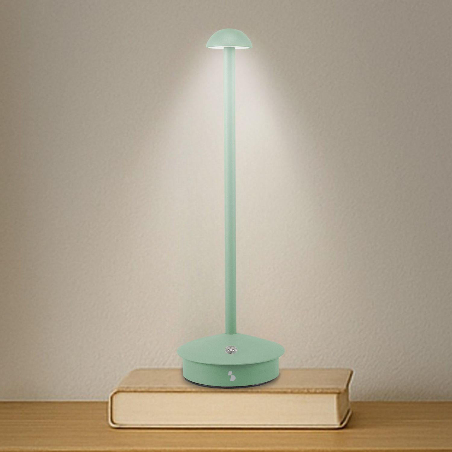 Una lampada da scrivania verde menta è su un libro. La lampada brilla. Lo sfondo è un muro beige e un tavolo di legno.